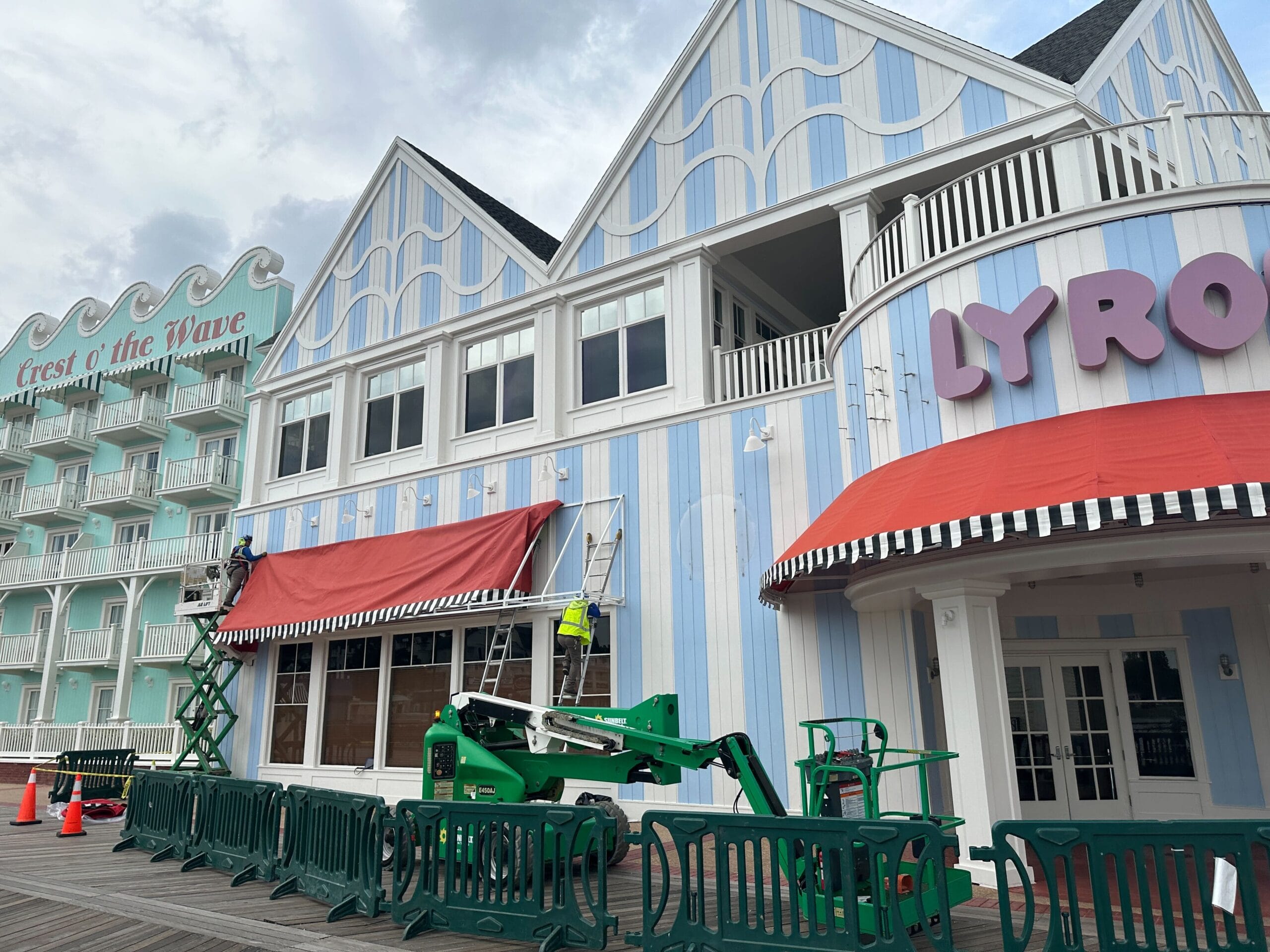 Two workers on lifts repaint a blue and white striped building with Jellyrolls Awnings, near Crest of the Wave hotel.