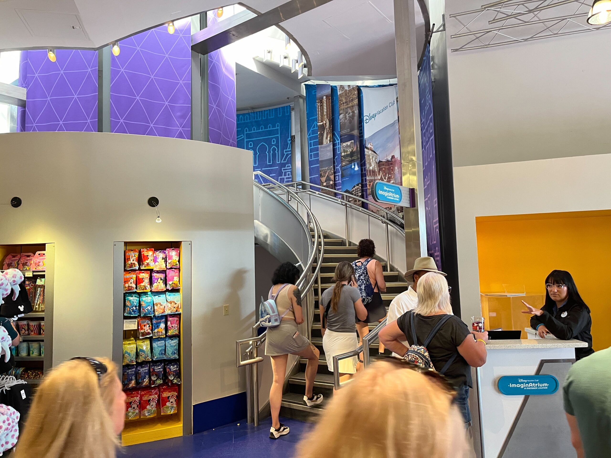 People ascending a spiral staircase in a brightly lit, refurbished store reminiscent of the sleek DVC Lounge at EPCOT, surrounded by colorful stands and a cashier stationed on the right.