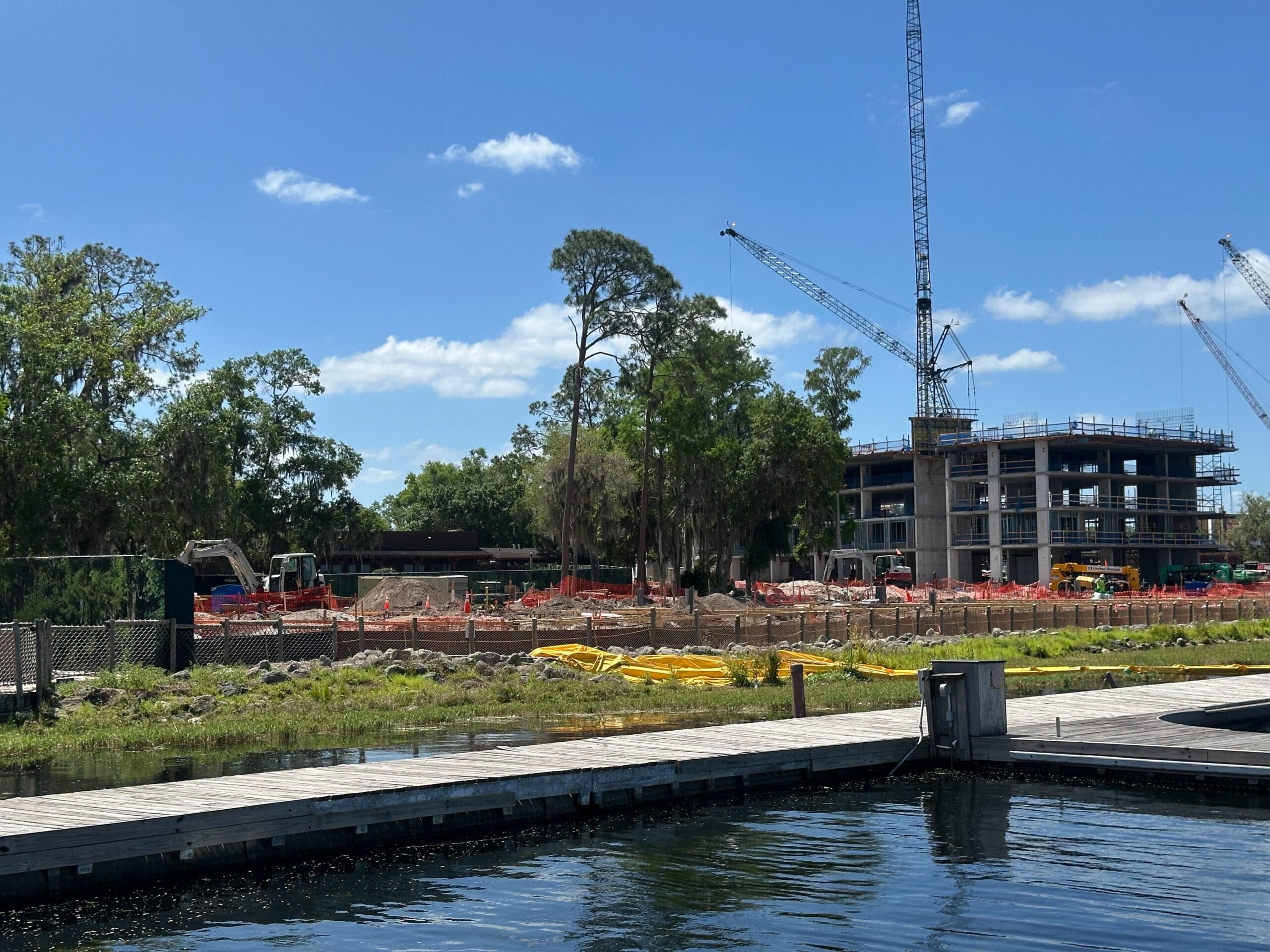 Construction site with cranes and partially built structures near a body of water. Trees and construction materials are visible, with a blue sky and scattered clouds overhead.