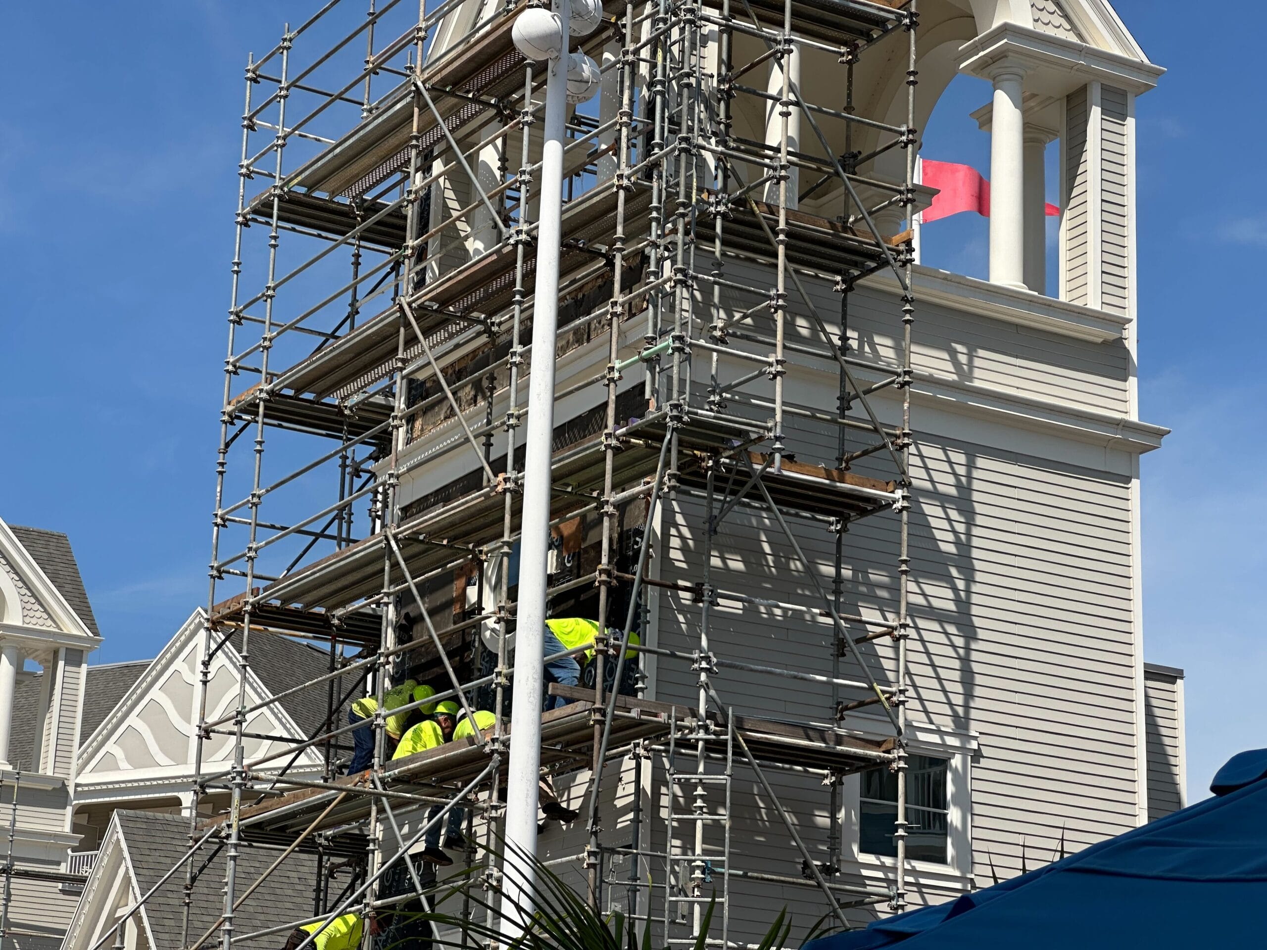 Workers in bright gear are on scaffolding set against a cream-colored building resembling Disney’s Yacht Club. A flag is visible atop the tower, adding a touch of charm to the scene.