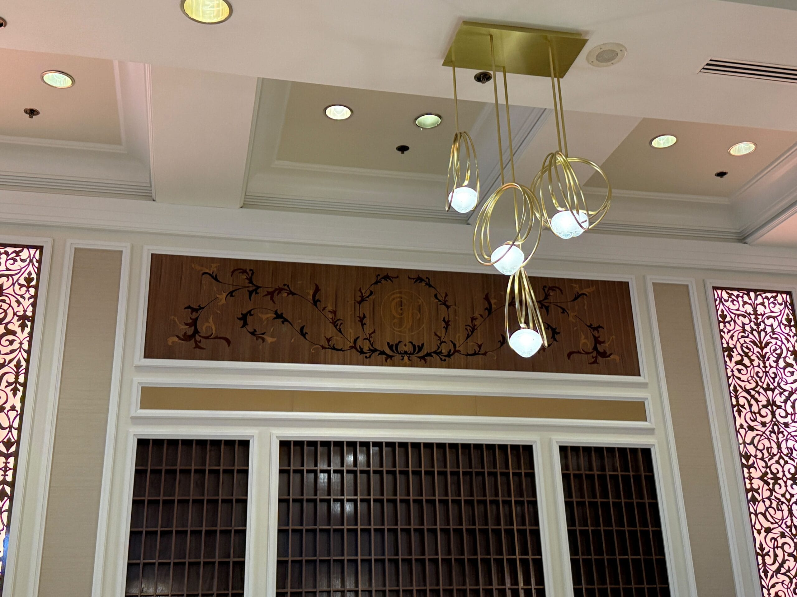 Modern gold light fixture hangs from a white ceiling, above a decorative wood panel and empty cubby shelves.