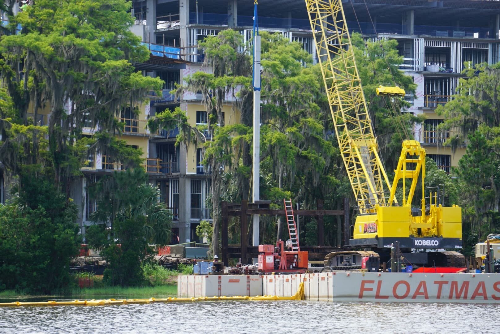 A yellow crane on a barge works near trees and a partially constructed building by the water.