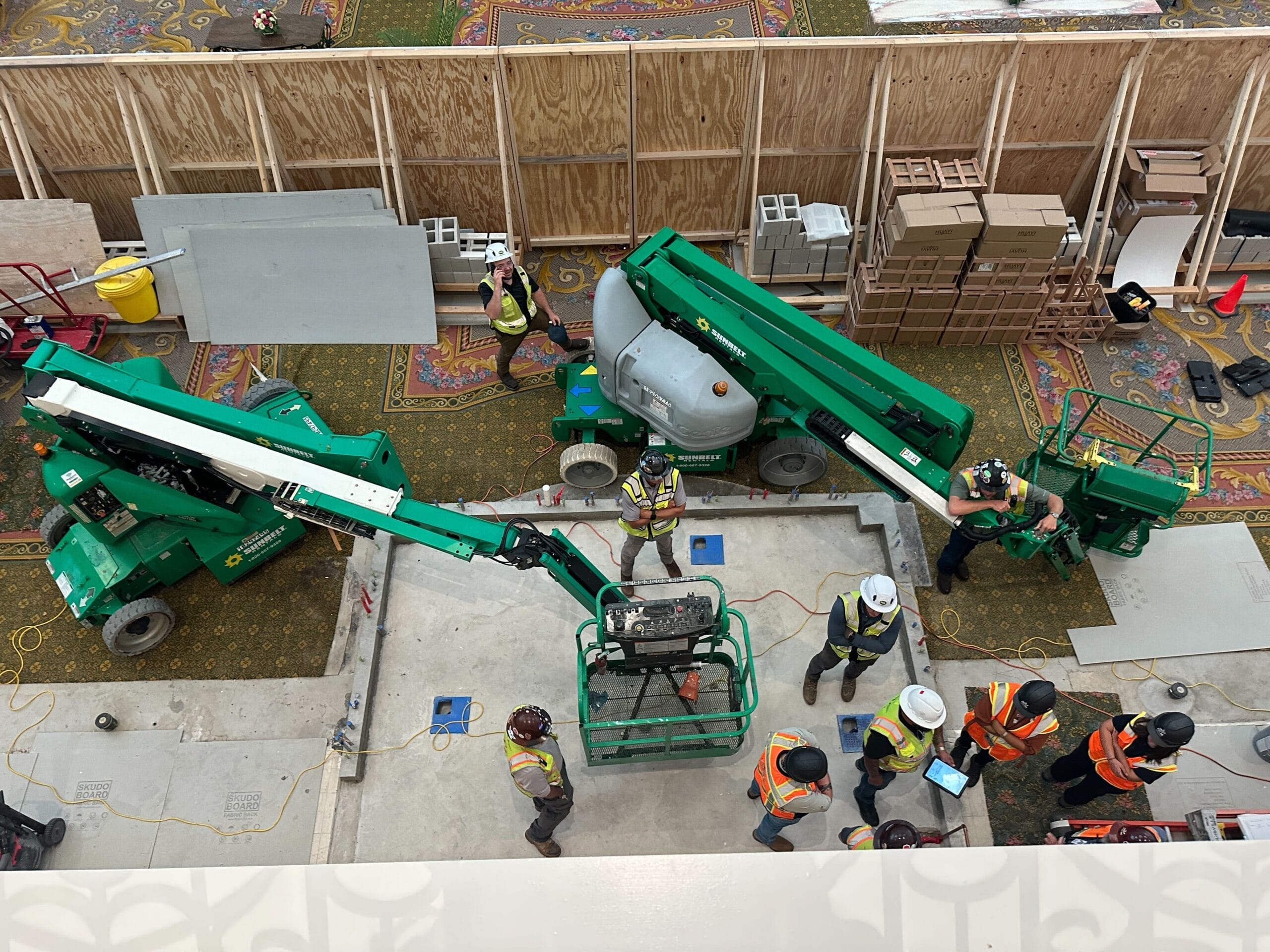 Construction workers in safety vests and helmets operate green lifts and equipment inside a large building under construction.