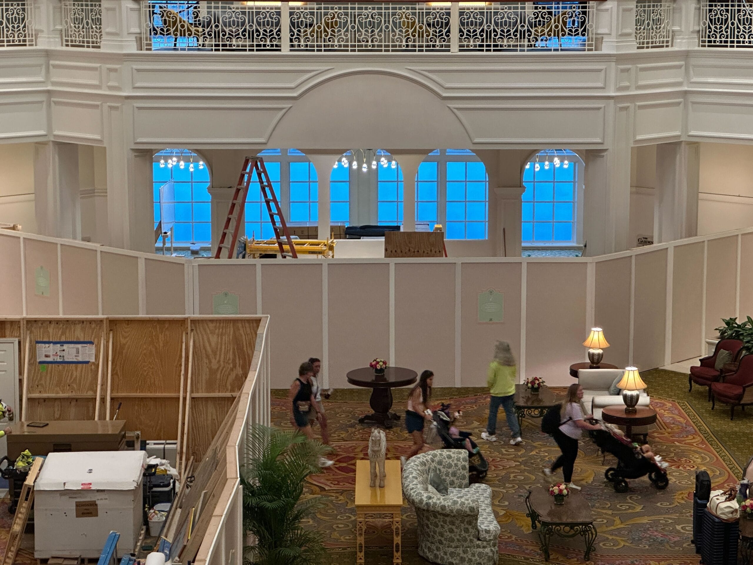 The Grand Floridian hotel lobby features construction barriers, people walking, and elegant furniture and lamps in the foreground.