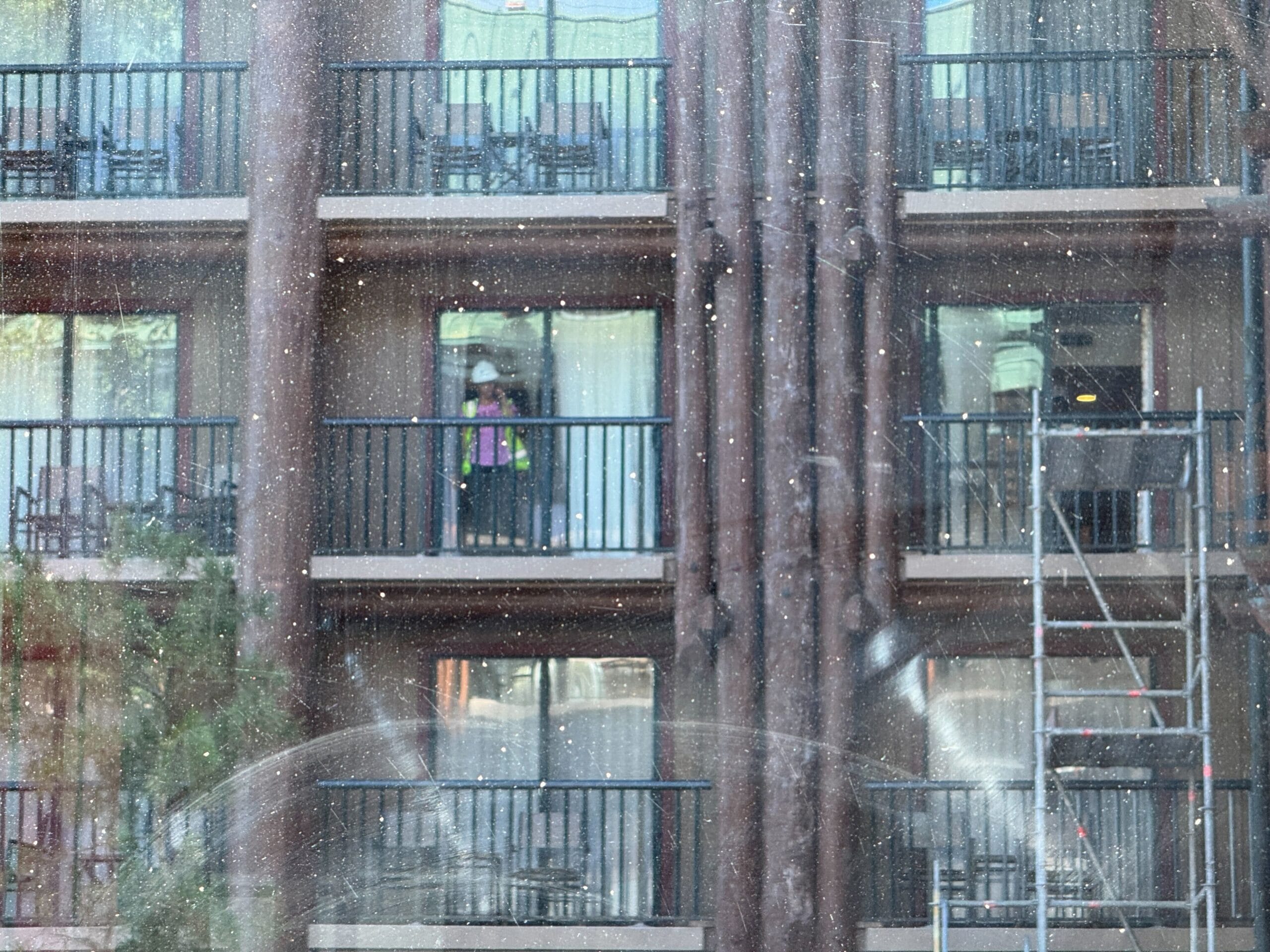 Person in a hard hat stands on a hotel balcony, seen through a dusty window, with scaffolding nearby.