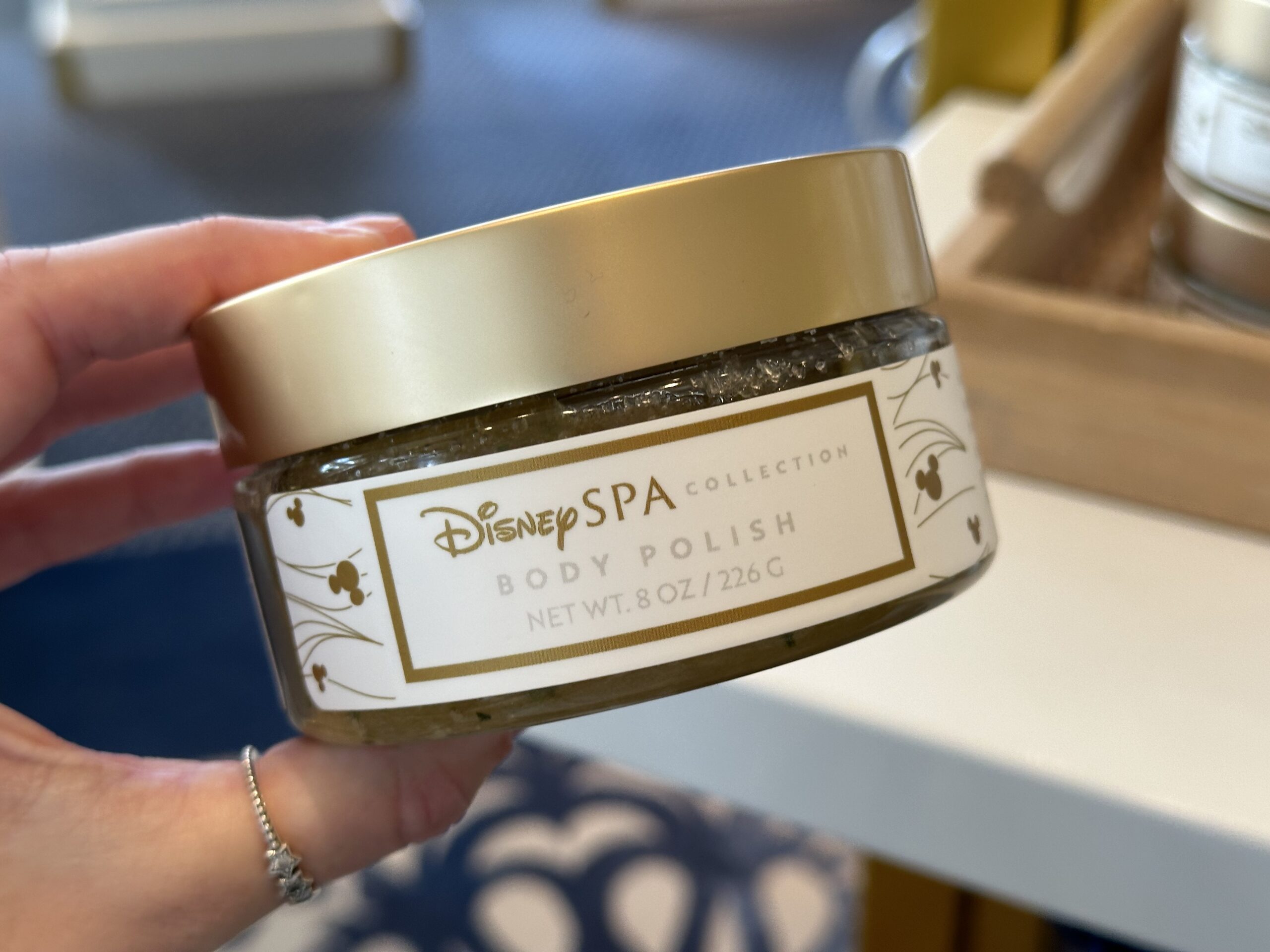 A hand holds a jar labeled Disney Spa Collection Body Polish with a net weight of 8 oz (226 g), reminiscent of the luxurious treatments at Disney World's Grand Floridian.