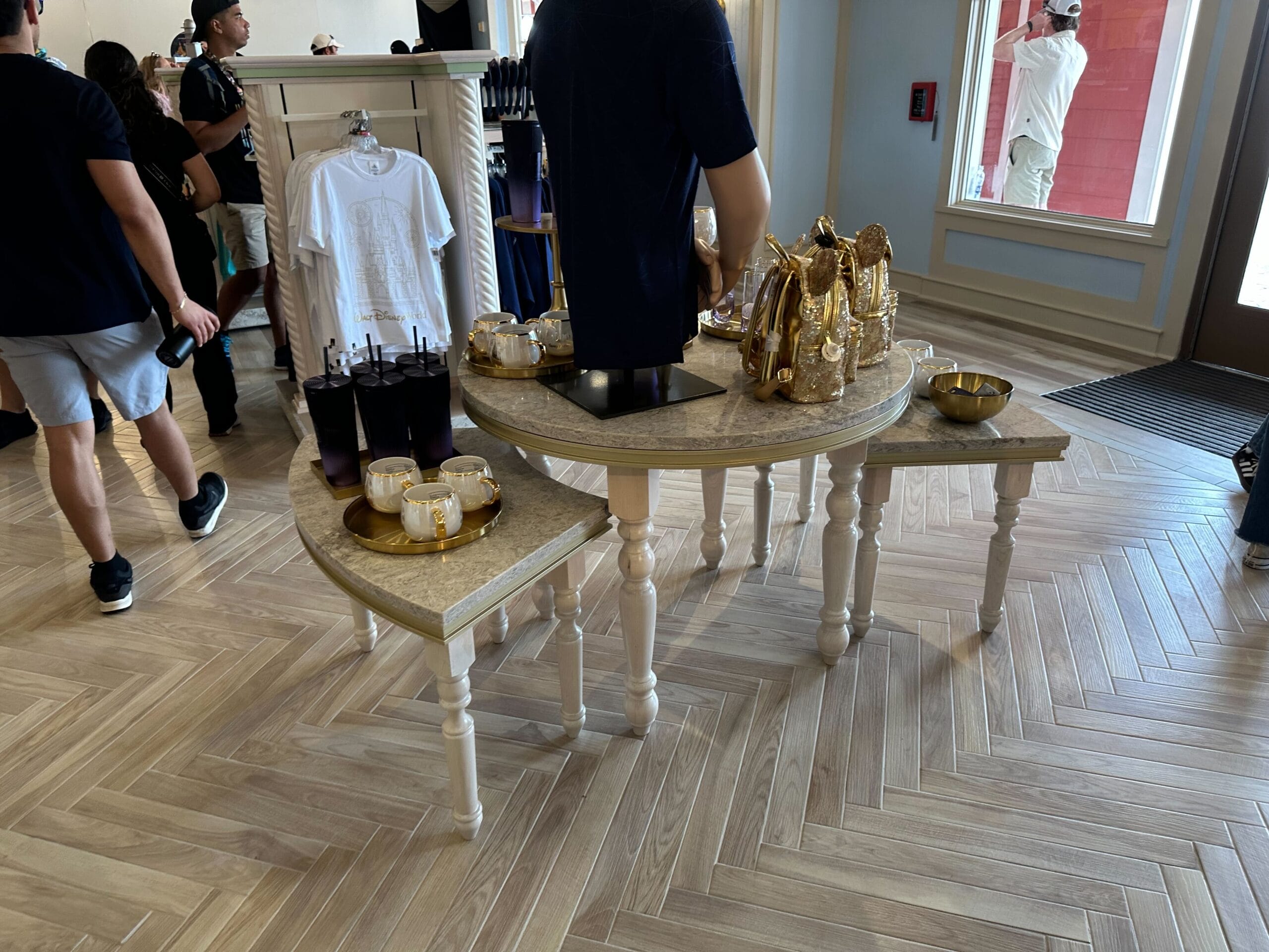 The display table at Screen Door General Store boasts an inviting array of drinks, snacks, and decorative items. Clothing options can be found in the background of this renovated section. Guests at Disney's Boardwalk Inn enjoy browsing nearby.