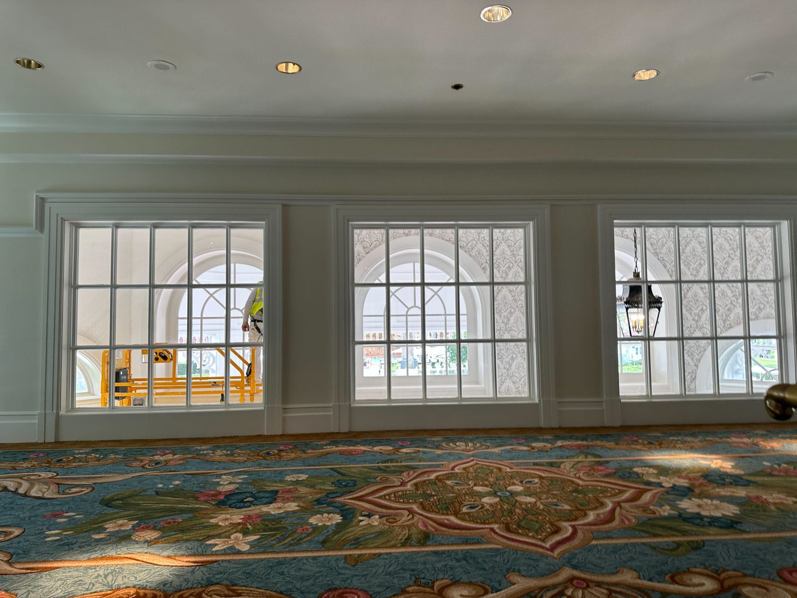 Three arched windows with white frames, ornate wallpaper, and a worker painting inside Disney's Grand Floridian Lobby—no castle in view.