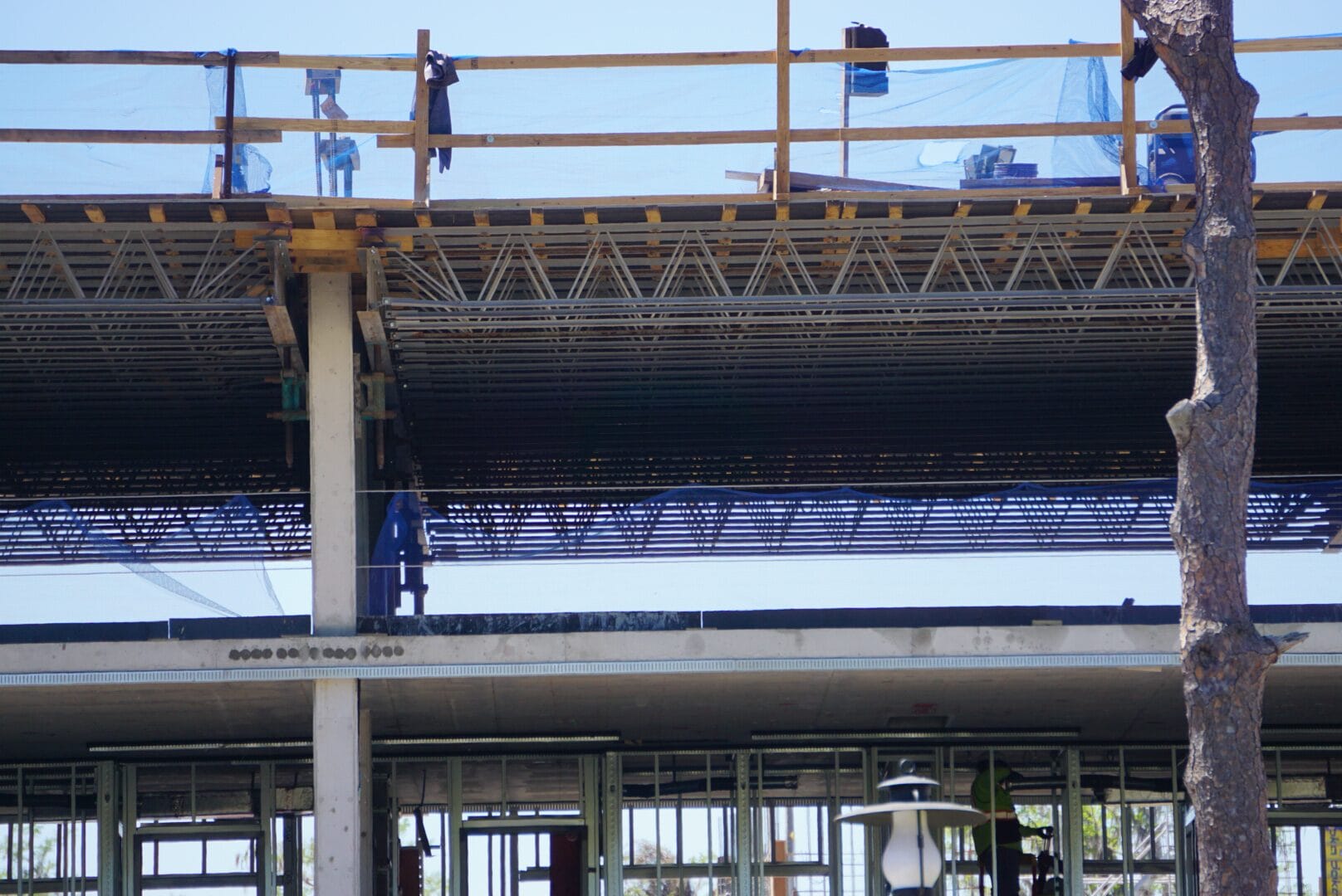 Construction site with a partially built multi-story structure. Visible metal beams, wooden scaffolding, safety netting, and tree in the foreground under a clear sky.
