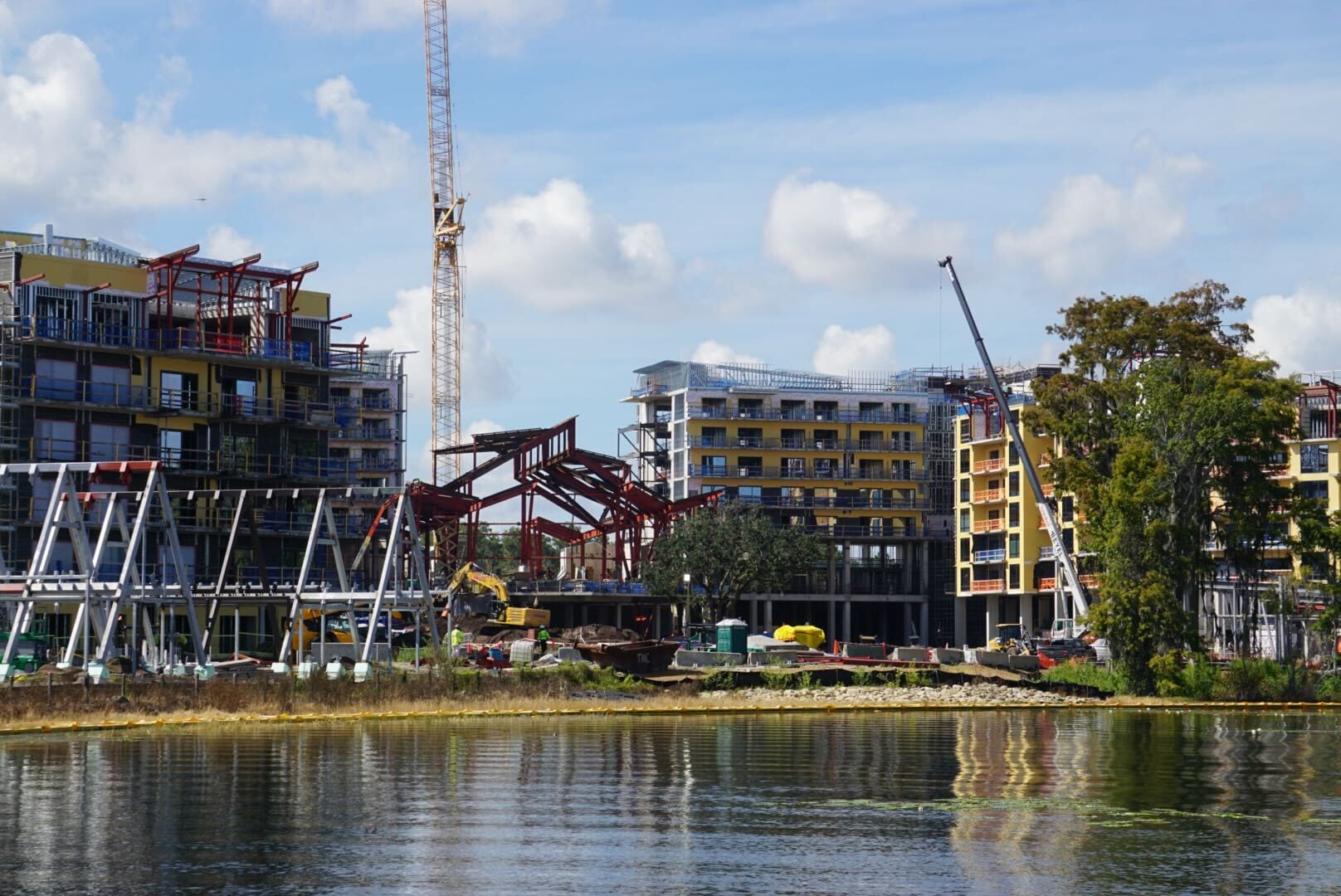 Construction site with cranes beside a pond, future home of Disney Lakeshore Lodge, under a blue sky with scattered clouds.