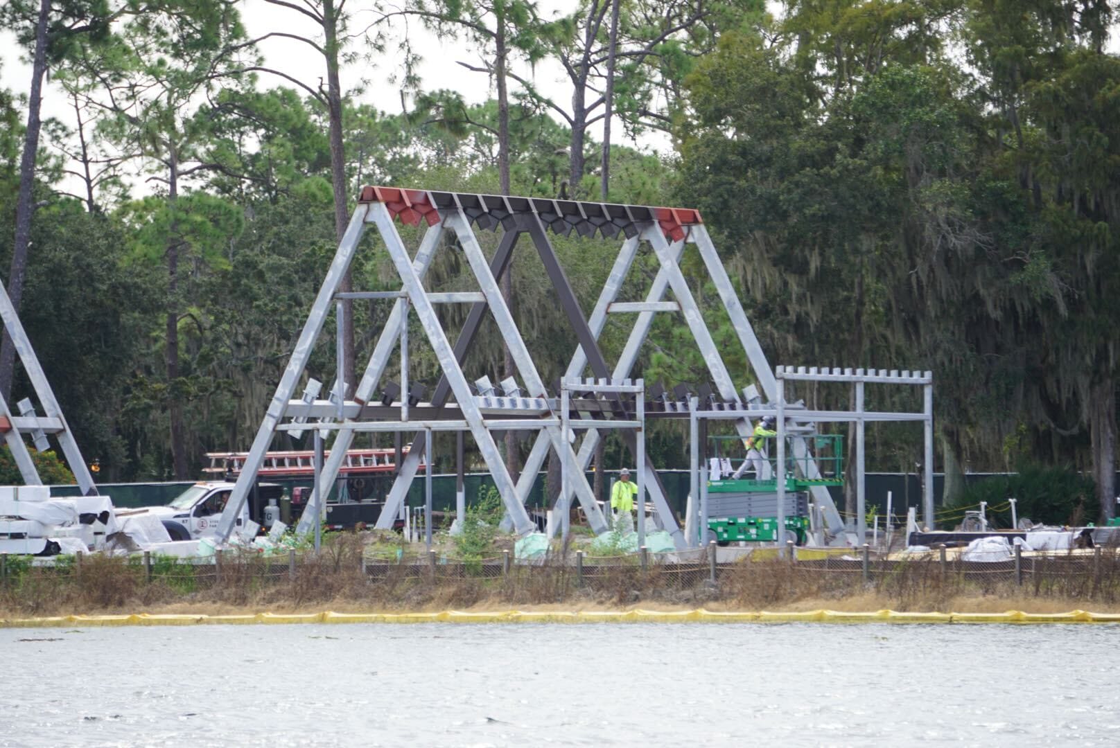 Construction Updates: Workers build a large A-frame metal structure at Lakeshore Lodge, near water and trees.