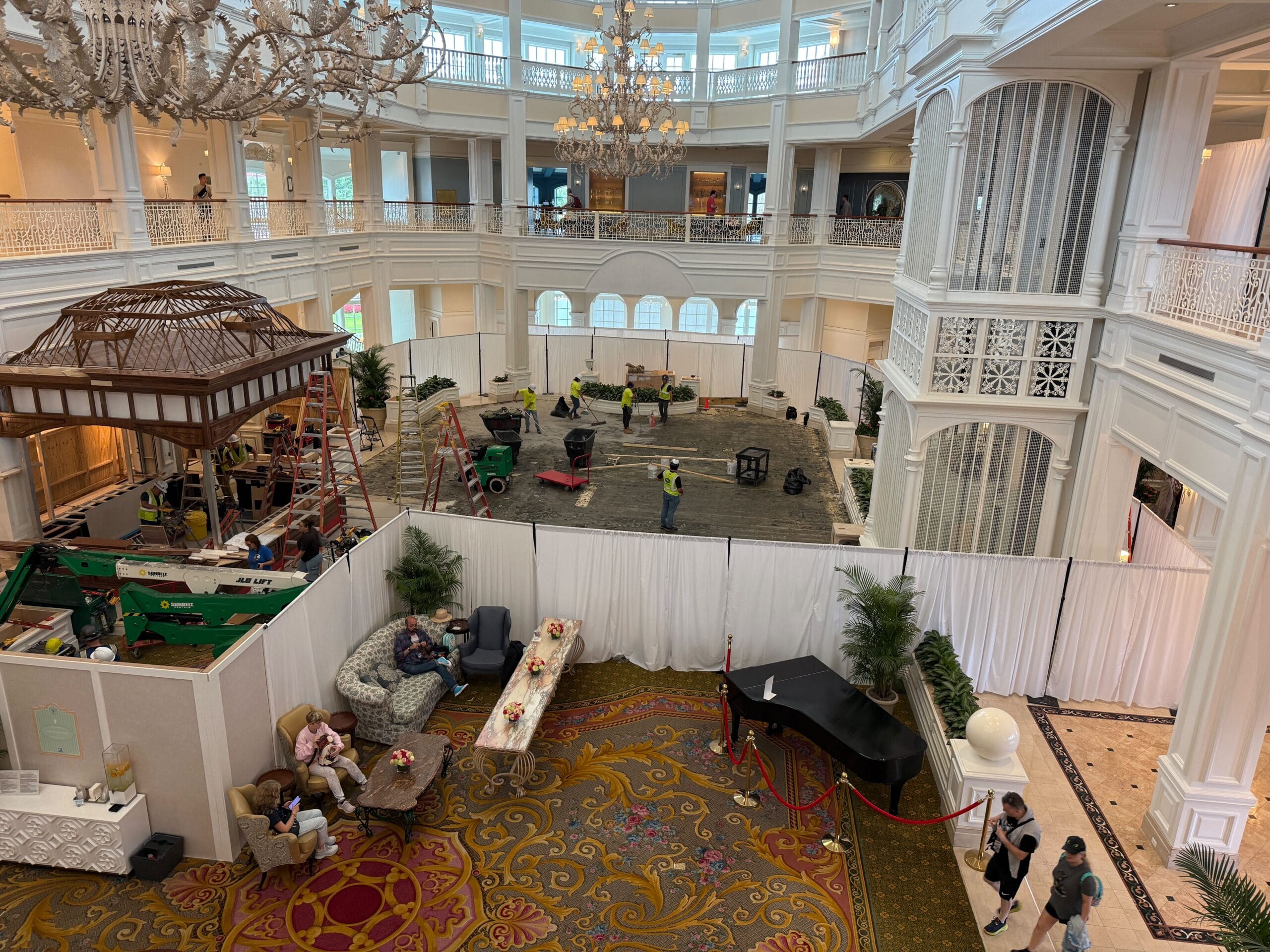 A hotel lobby under construction, with workers, equipment, and seating areas; guests relax near a black piano.