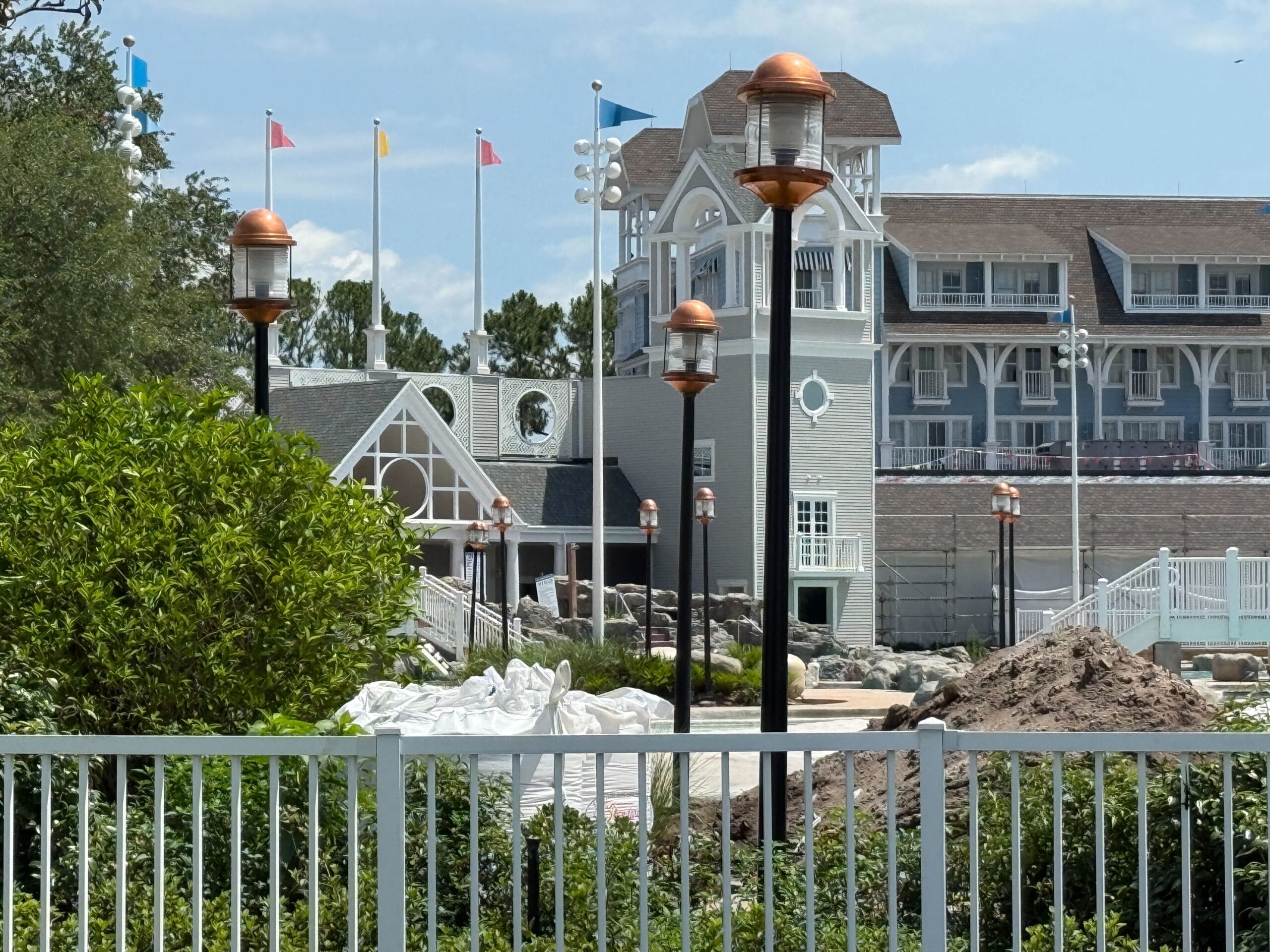 A construction area near a gray building with white trim, lampposts, flags, and green bushes under a blue sky.