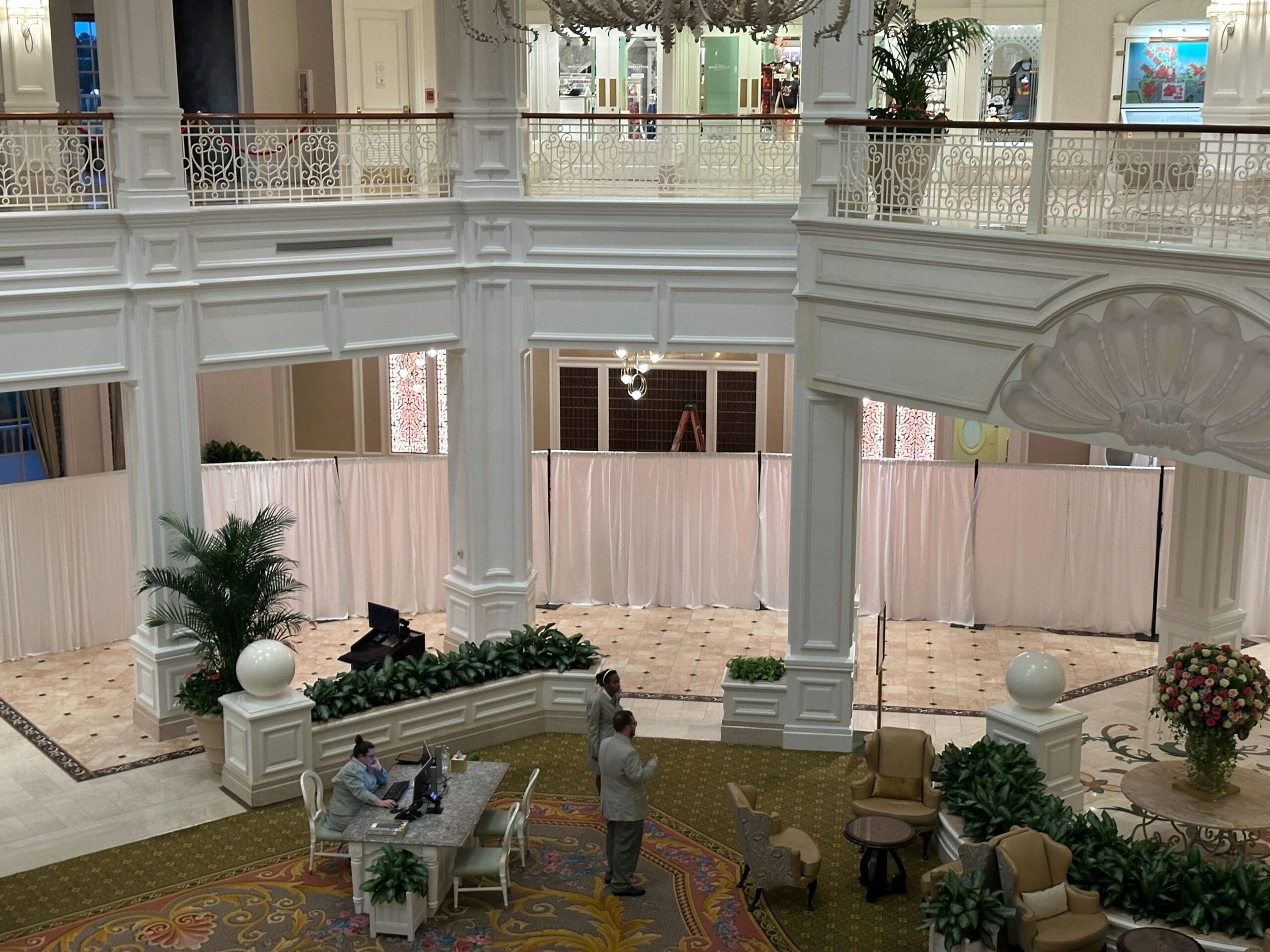 Two people stand talking in a grand hotel lobby near a white divider curtain, surrounded by plants and elegant decor.