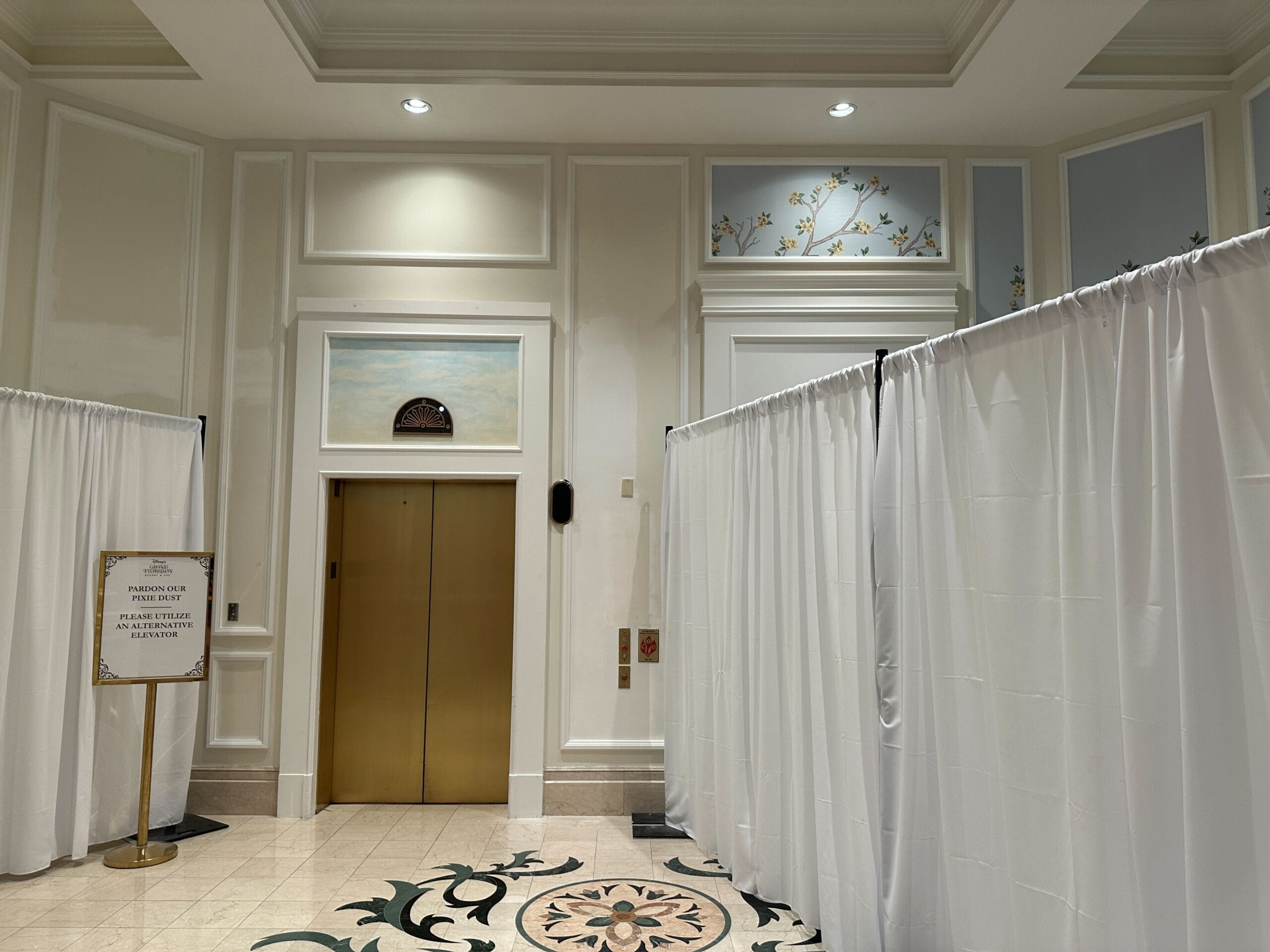 A gold elevator is surrounded by white privacy curtains and a sign on a stand in a decorative hotel hallway.