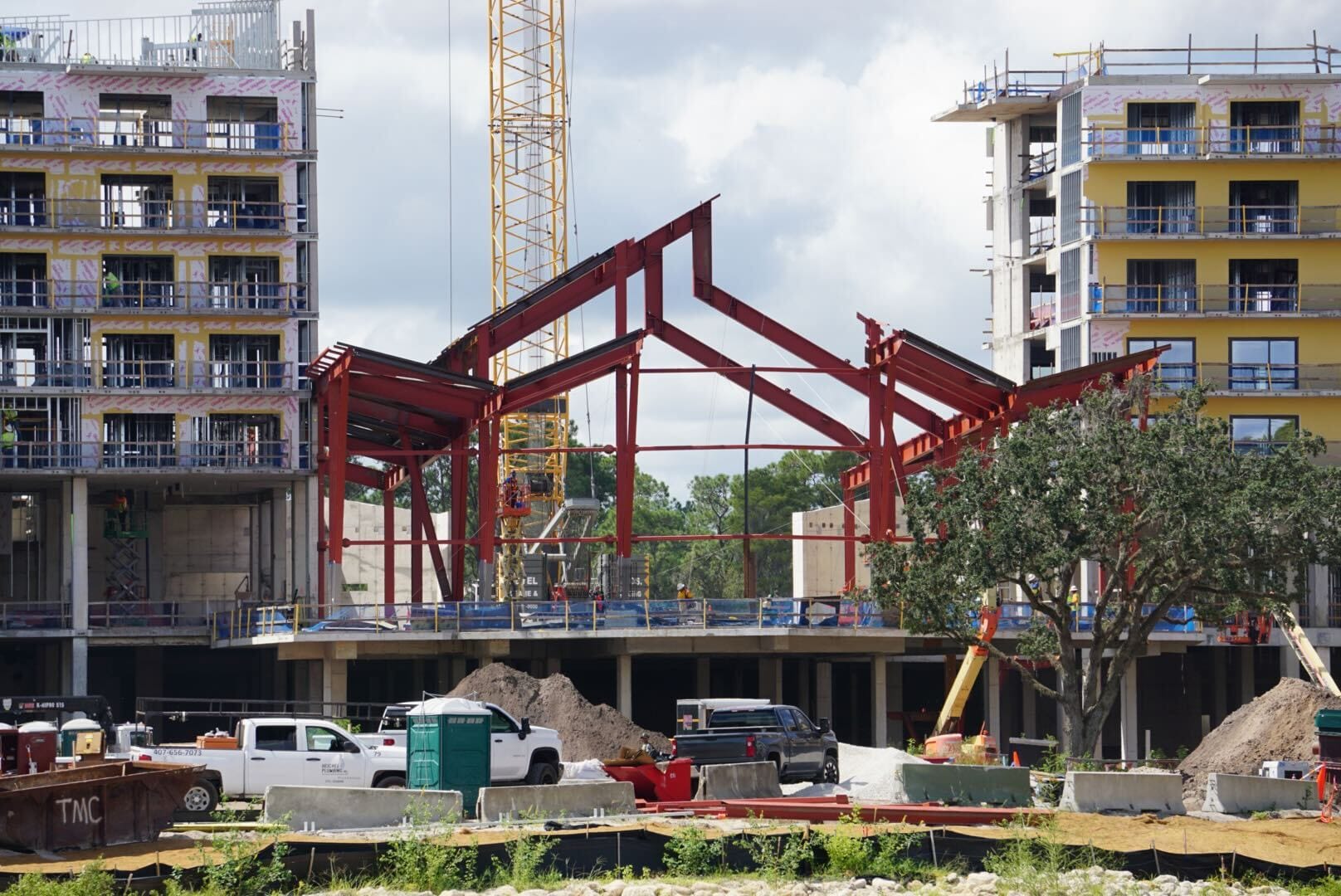 Steel framework of a building under construction between two partially completed structures, with vehicles and equipment nearby.