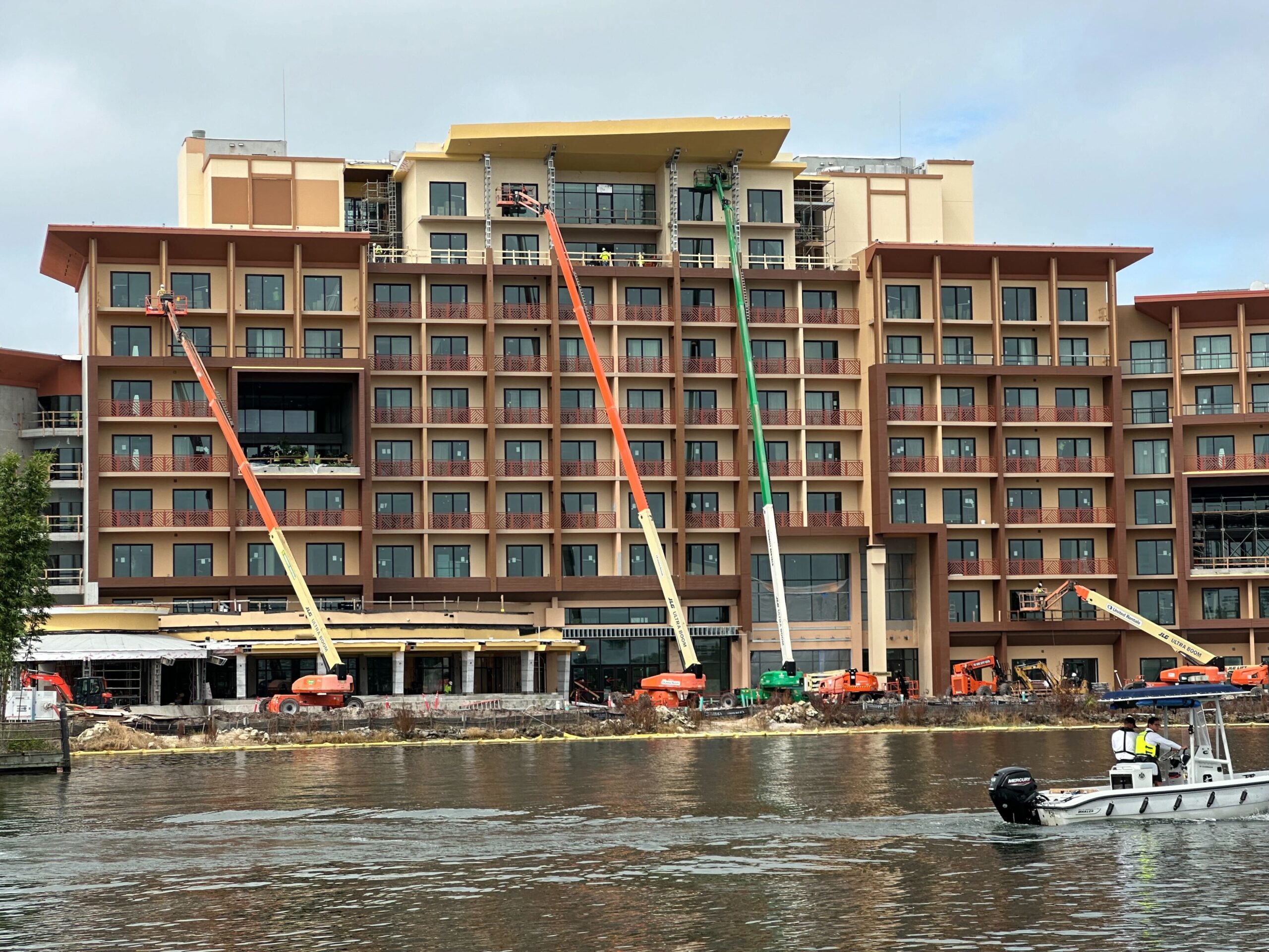 Multiple cranes are being used for construction work on a multi-story building by a waterfront, with a boat passing by in the foreground.
