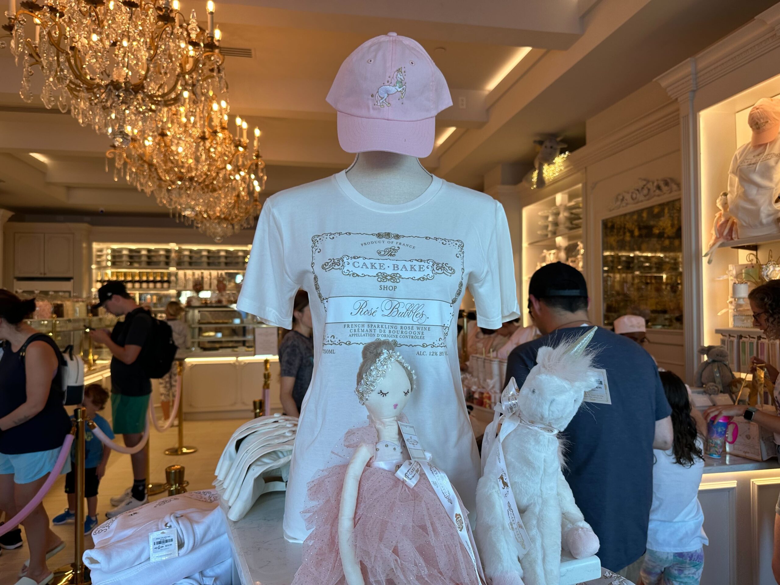 Mannequin wearing a T-shirt and cap in a boutique. Chandelier overhead, people browsing, and decorative display with plush dolls in the foreground.