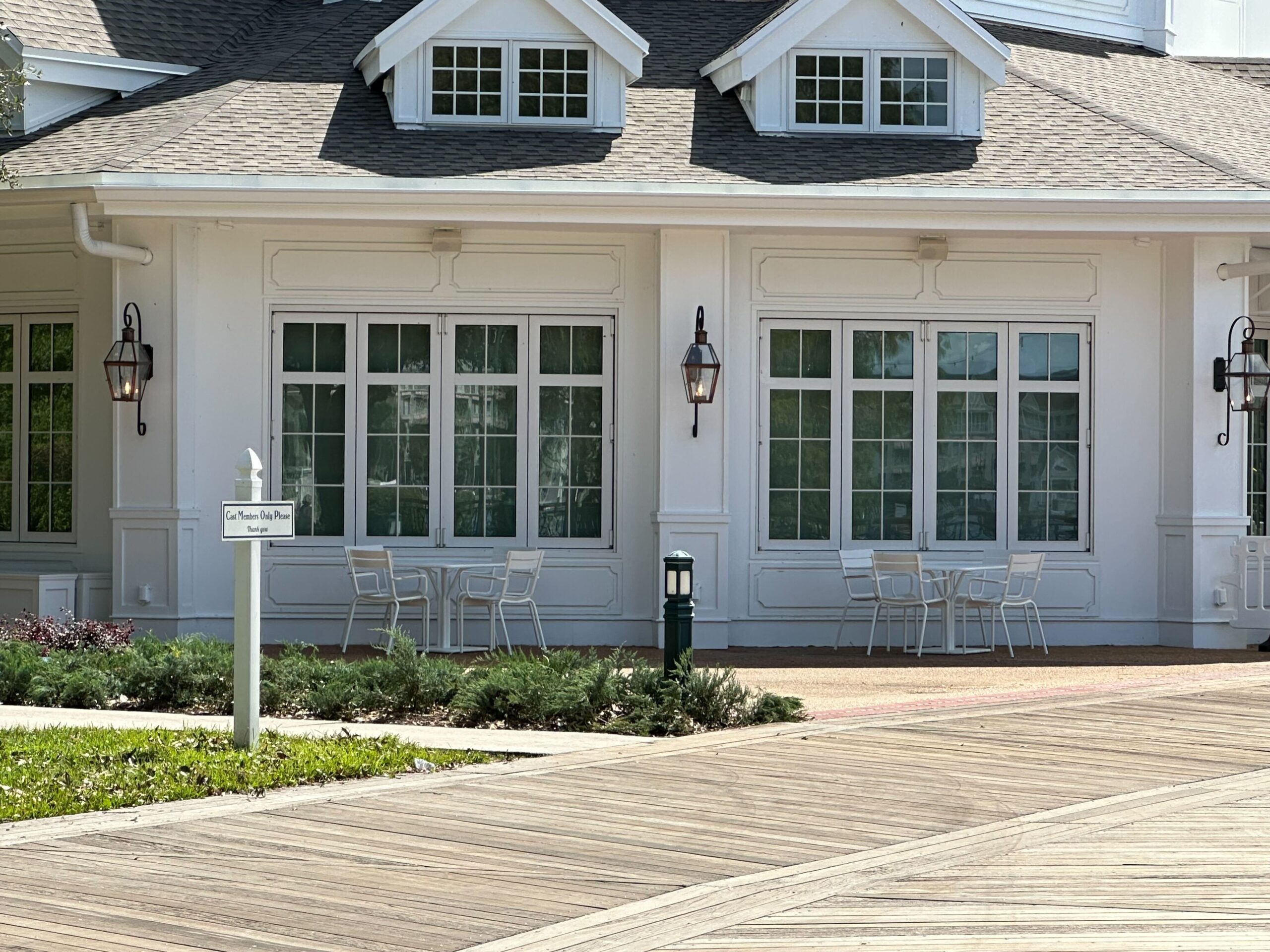 A building with white siding and large windows, the Cake Bake Shop boasts outdoor tables and chairs on a wooden deck reminiscent of Disney's BoardWalk. A sign is visible in the foreground.