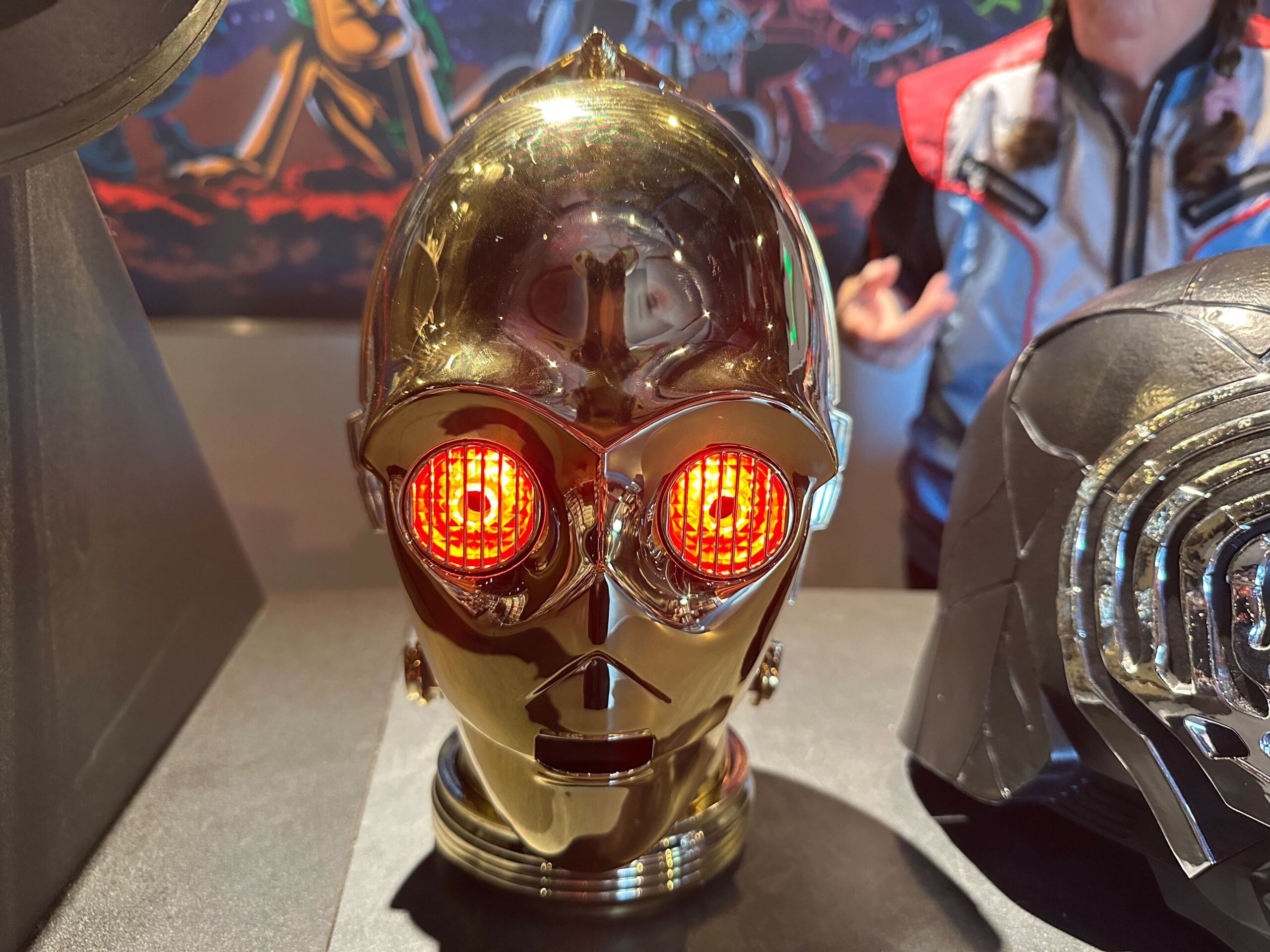 A golden C-3PO premium electronic head with red glowing eyes, reminiscent of Star Wars droids, sits on a table next to a black helmet. A person in a jacket is partially visible in the background, evoking scenes you'd expect to find at Disney World.