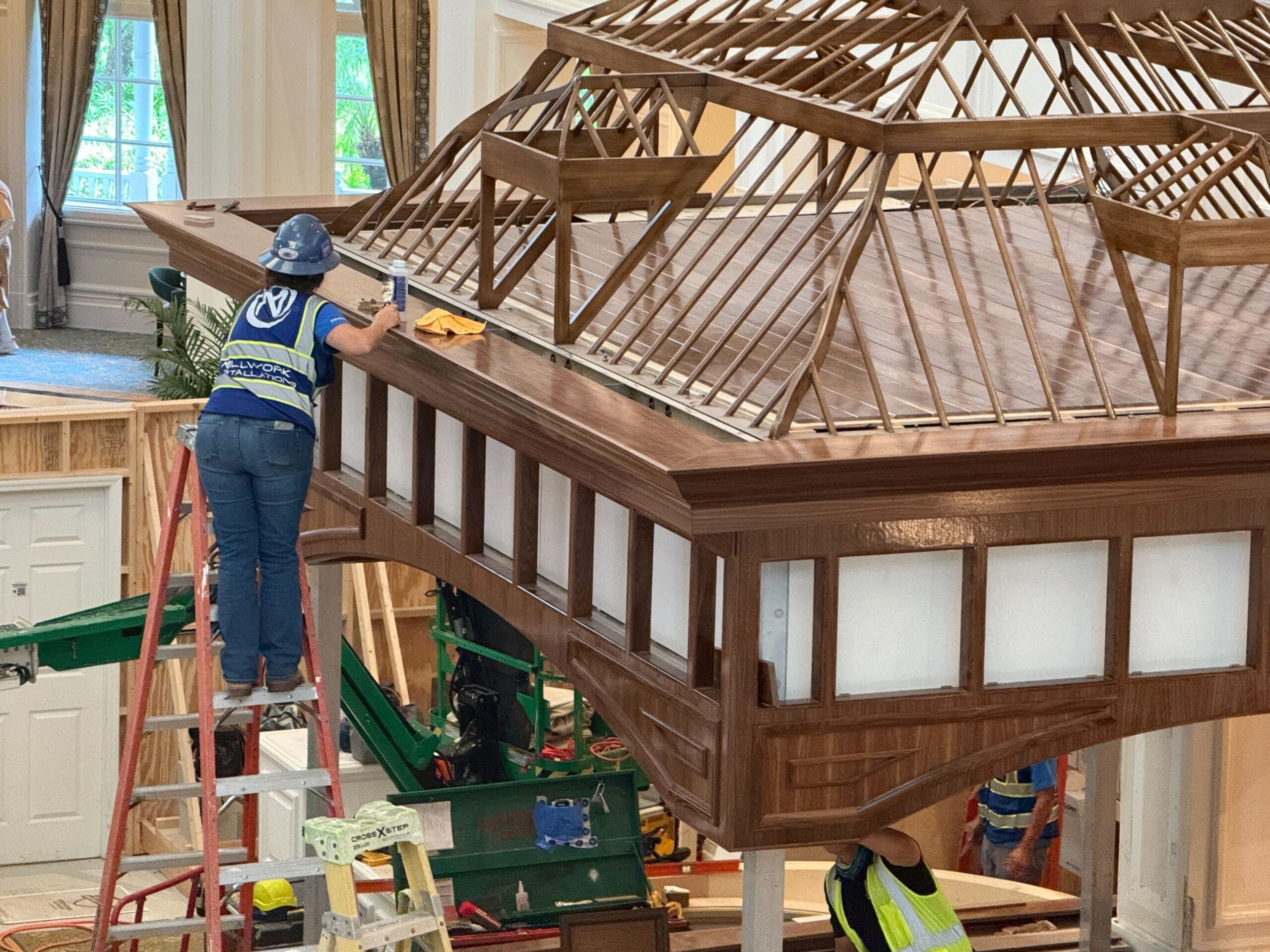A worker stands on a ladder cleaning a large wooden architectural structure indoors, while another worker stands below.