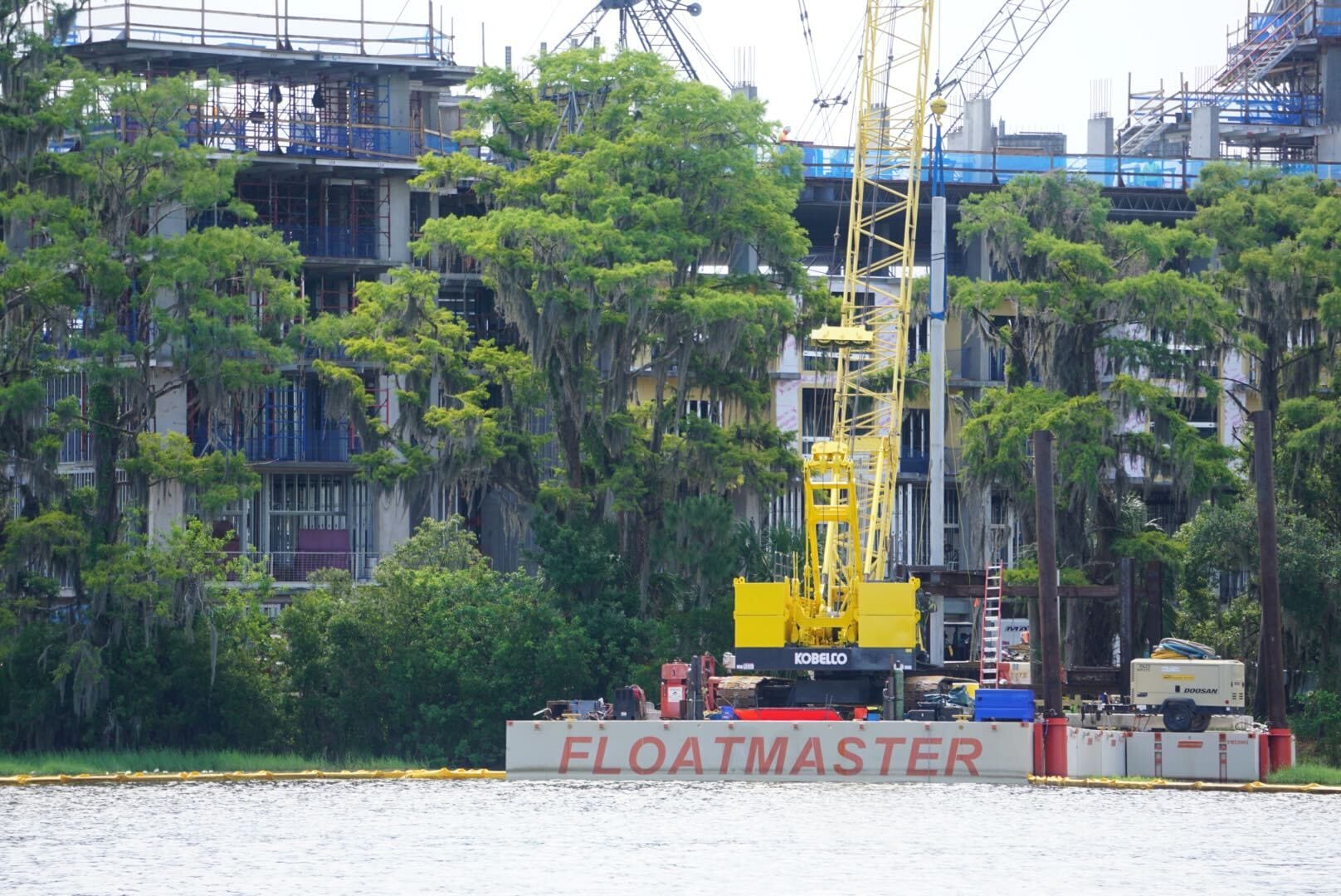 Yellow crane on FLOATMASTER barge near waterfront, with trees and a building under construction in the background.