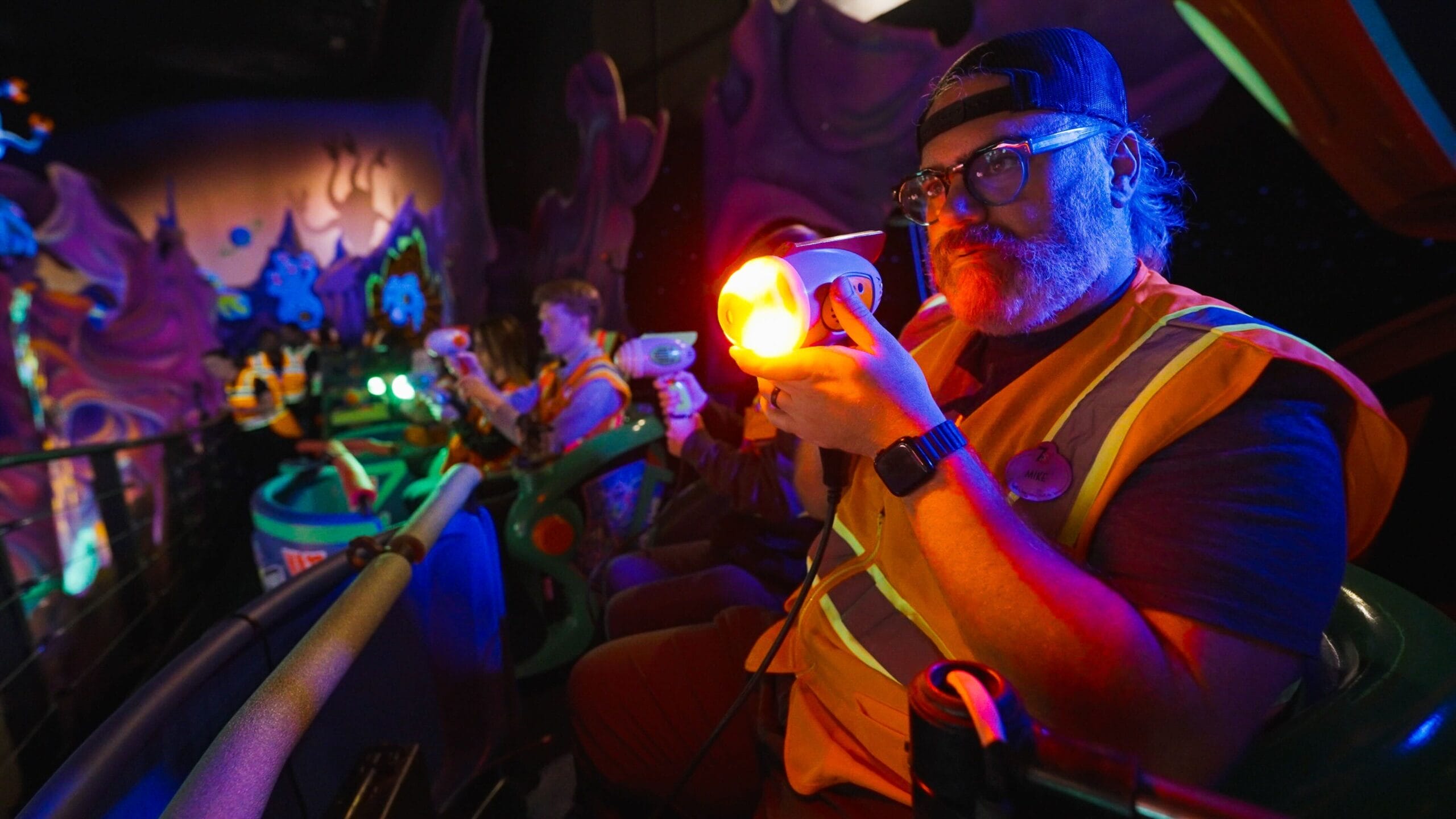 A person wearing a cap and safety vest uses a handheld light device on the Buzz Lightyear's Space Ranger Spin ride in Magic Kingdom. The colorful, interactive environment is filled with other visitors enjoying the experience in the background.