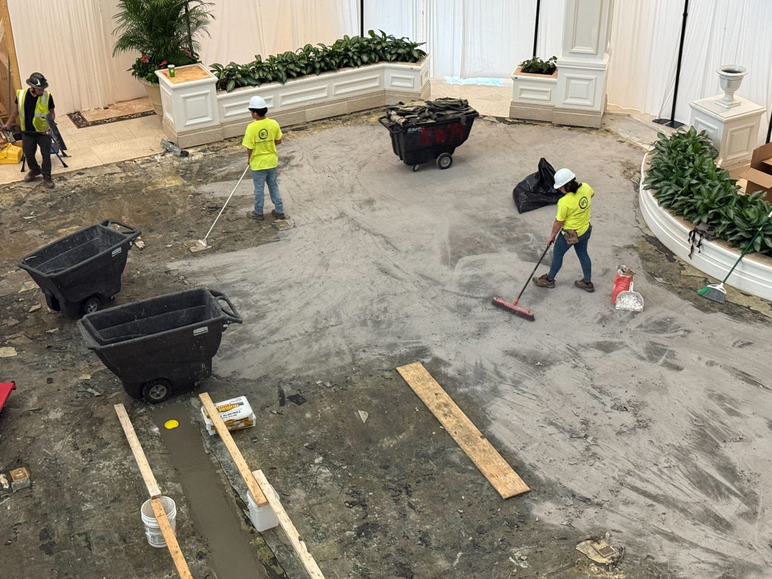 Three construction workers sweep and clean a dusty indoor worksite surrounded by planters and white drapes.