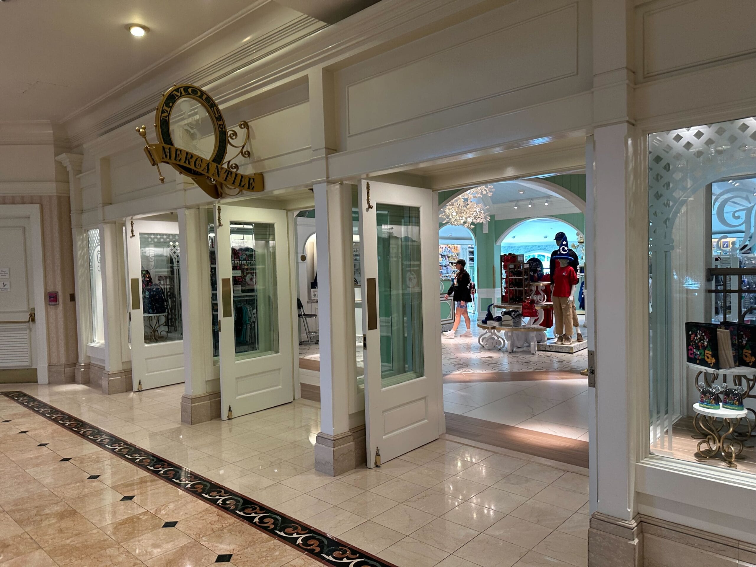Entrance to the boutique store Acadiana Mercantile with open double doors reveals a Grand Floridian-inspired interior. Mannequins display clothing and accessories, while the tiled flooring with decorative inlays adds an artistic touch.