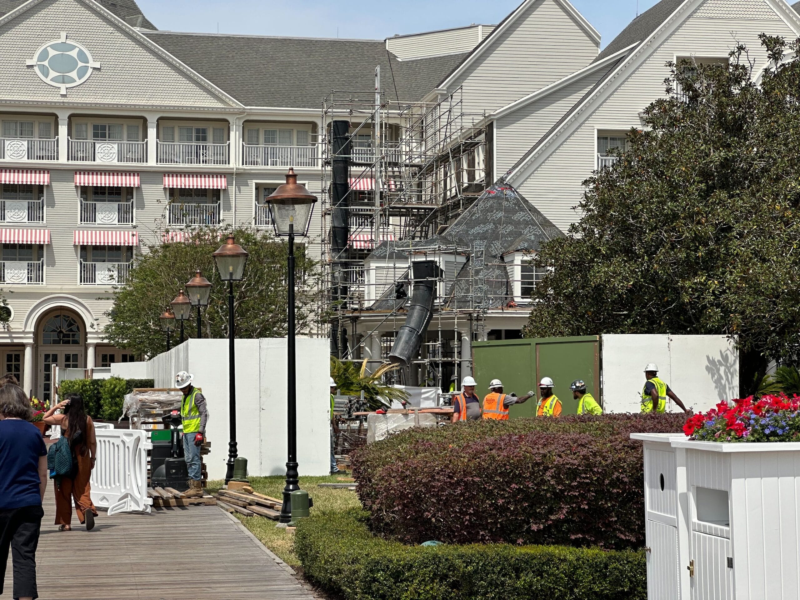 Construction workers in safety gear stand near scaffolding at Disney's Yacht Club, where a building with striped awnings is under renovation. The area, featuring pool construction, is partially enclosed by white barriers, while plants and flowers bloom in the foreground.