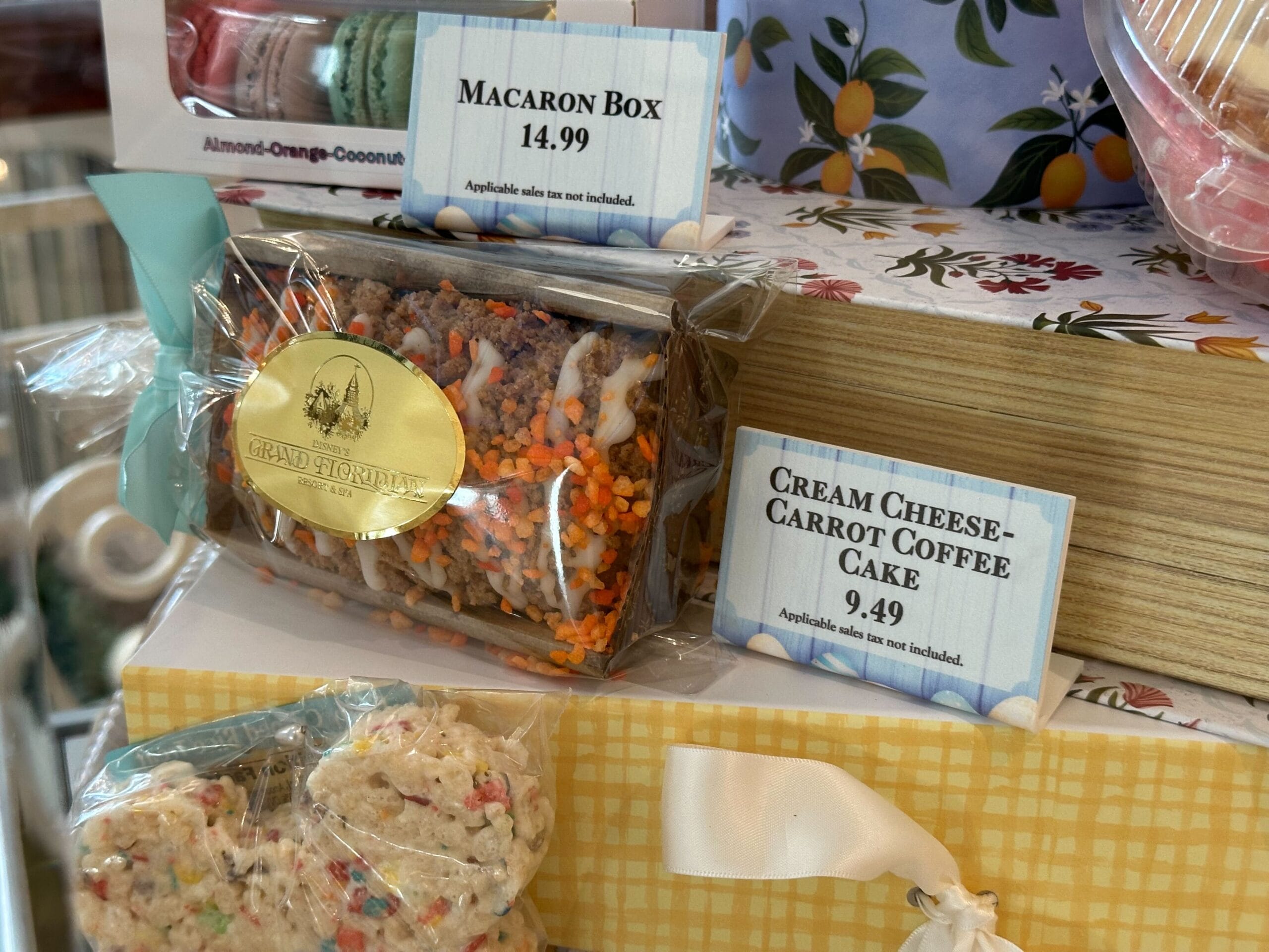 Packaged macaron box priced at $14.99 and cream cheese carrot coffee cake at $9.49 displayed on a floral-patterned counter.