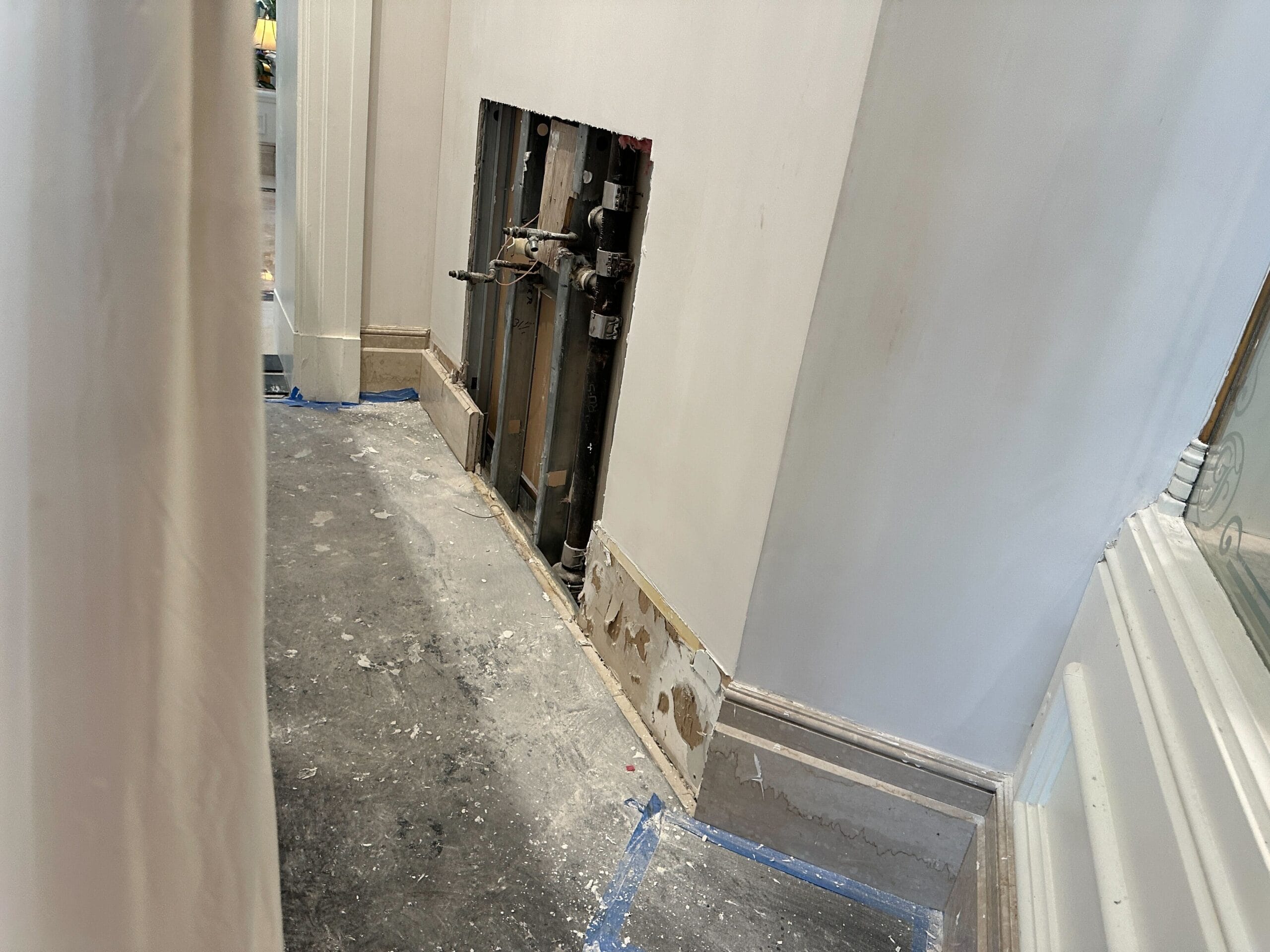 Wall with a rectangular section removed, exposing pipes; dust and debris cover the tile flooring from construction work.