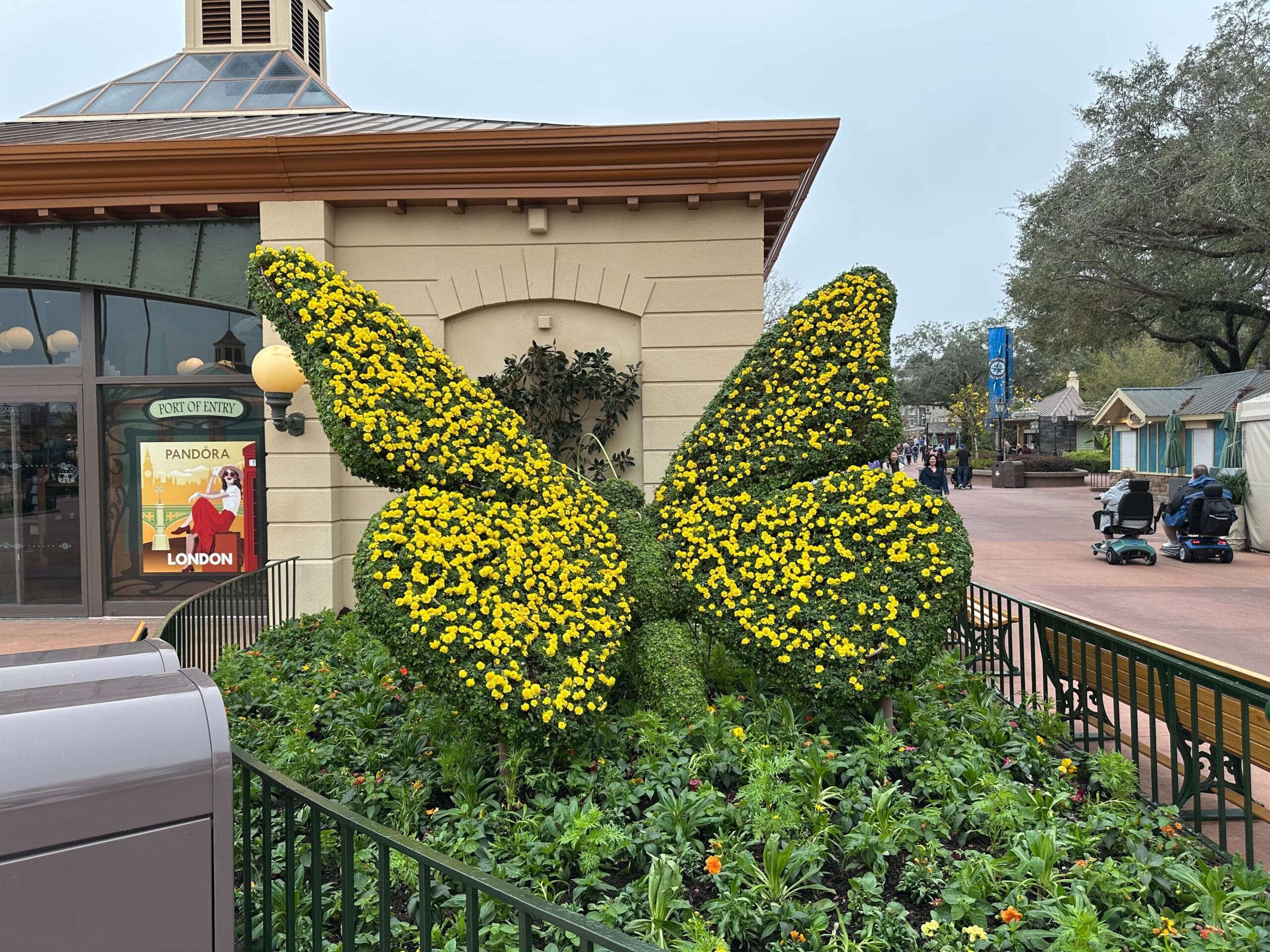 A butterfly-shaped topiary covered in yellow flowers stands in a landscaped area next to a building with a poster and a street in the background.