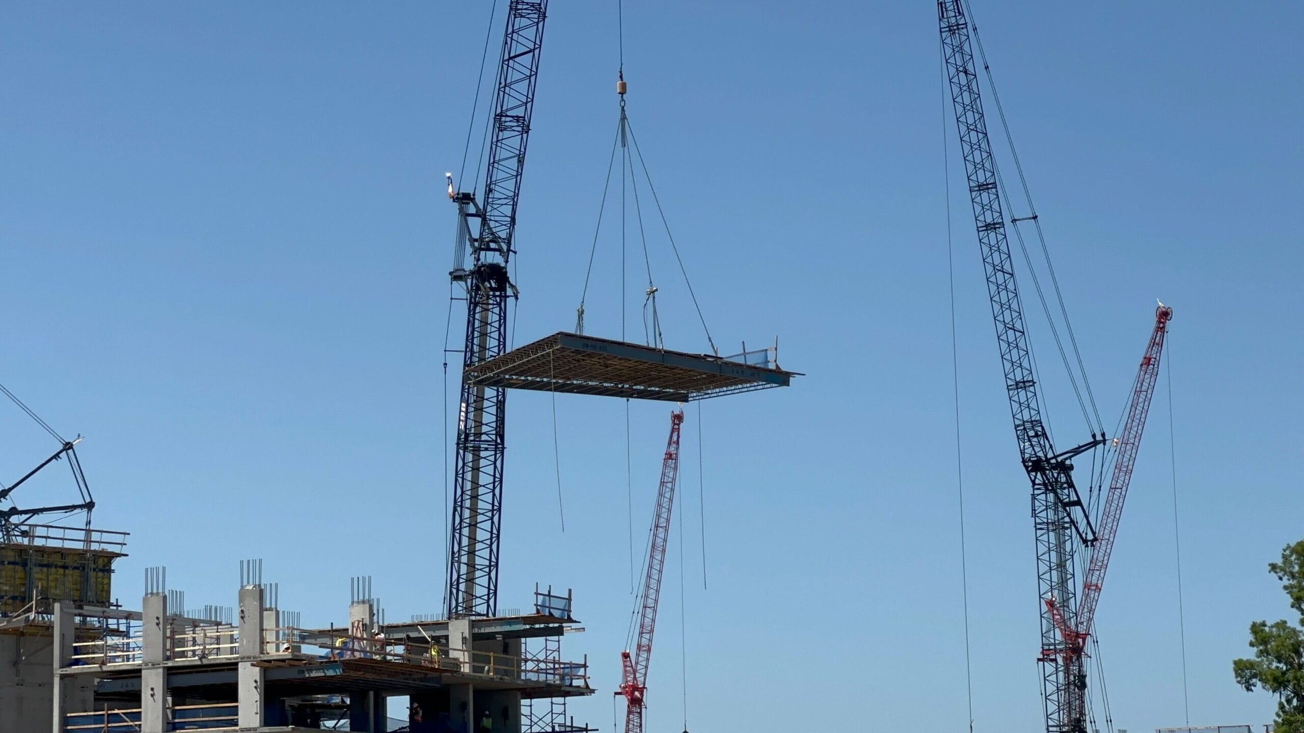 Two cranes lift a large steel frame at a construction site near Lakeshore Lodge. A partially constructed building is visible in the background under a clear blue sky.