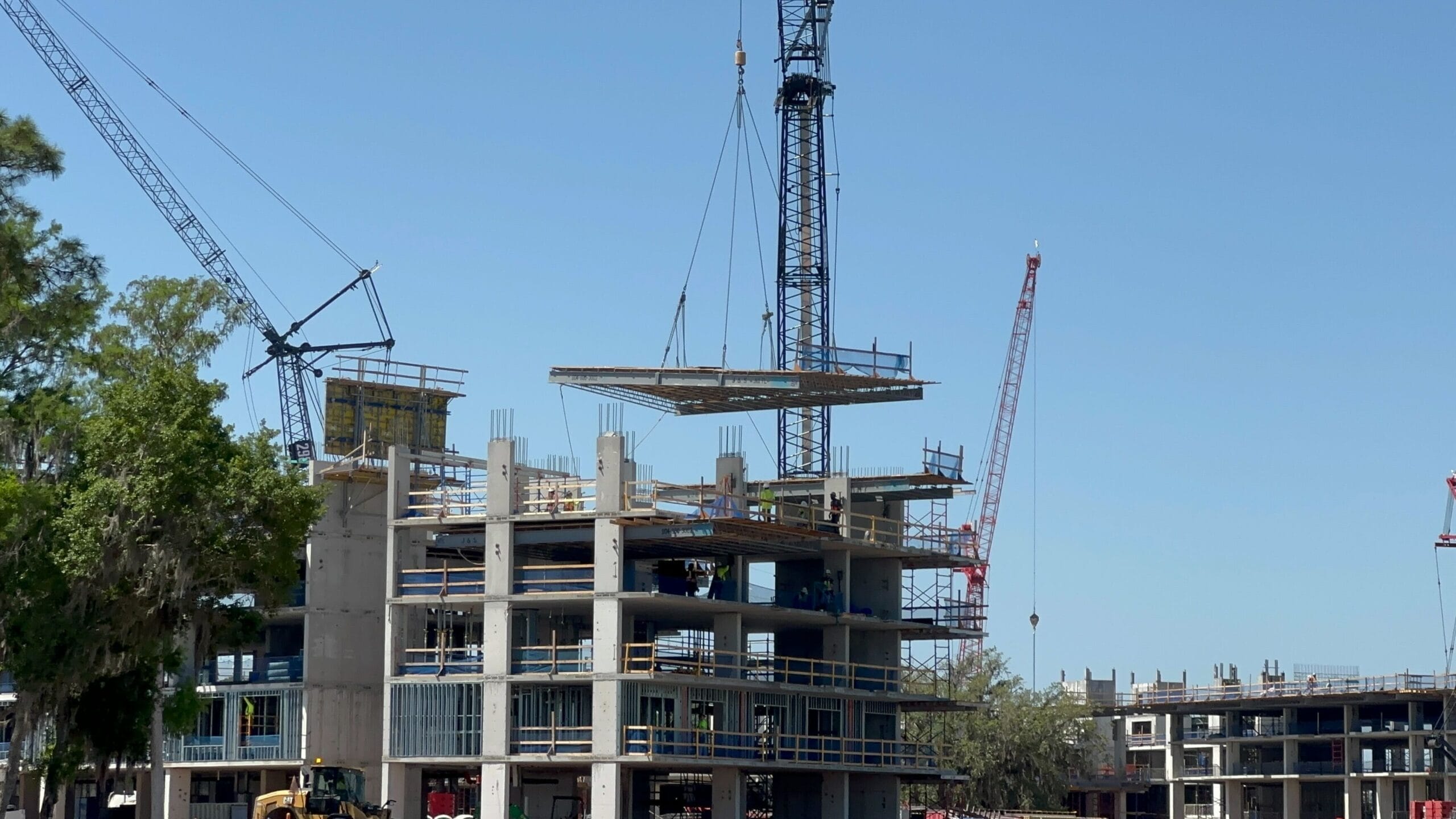 A crane lifts a large panel onto a multi-story building under construction near the serene lakeshore lodge. Additional cranes and structures are visible in the background under a clear blue sky.