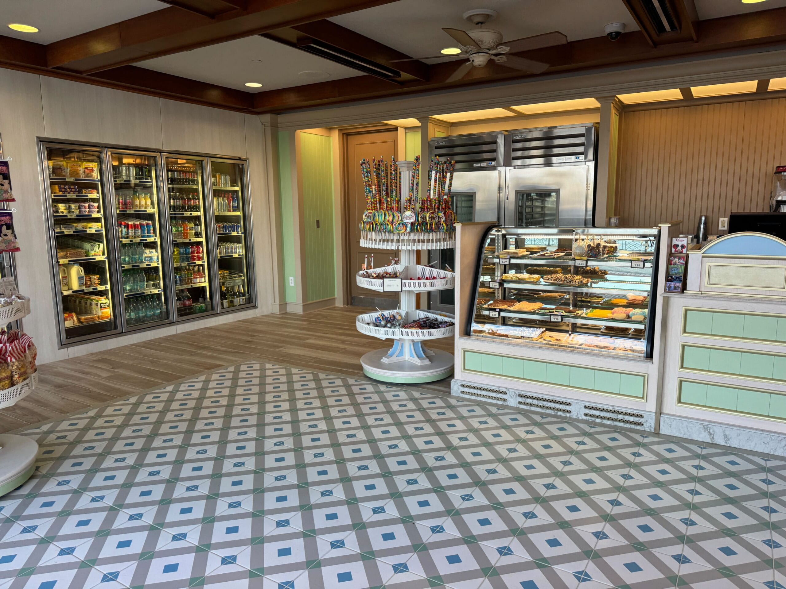 A bakery interior with display cases of pastries and a stand of colorful lollipops. Refrigerated drinks are visible on the left. The floor has a geometric tile pattern.