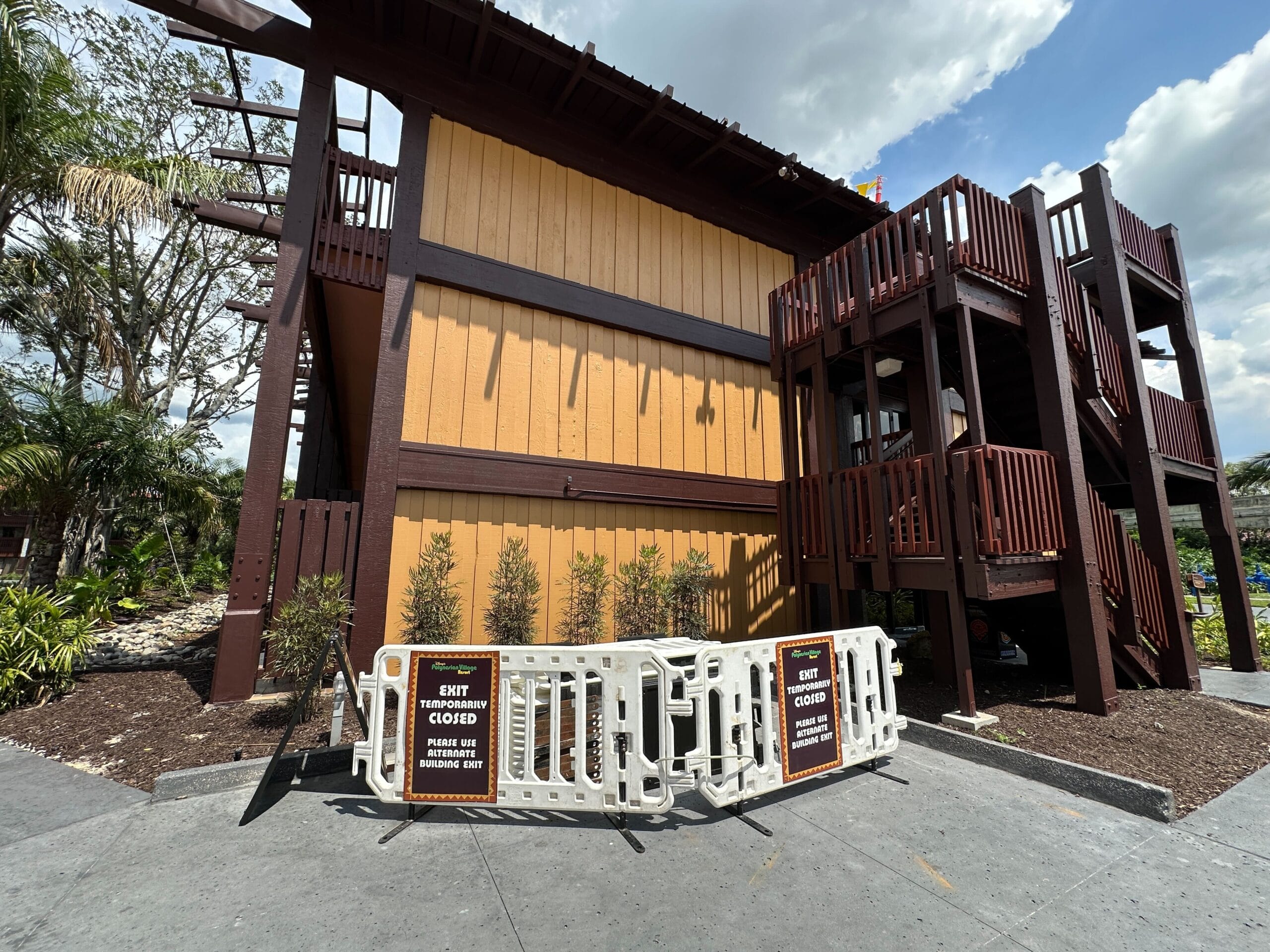 Two signs on barriers block a staircase exit outside a brown and yellow building under a partly cloudy sky.