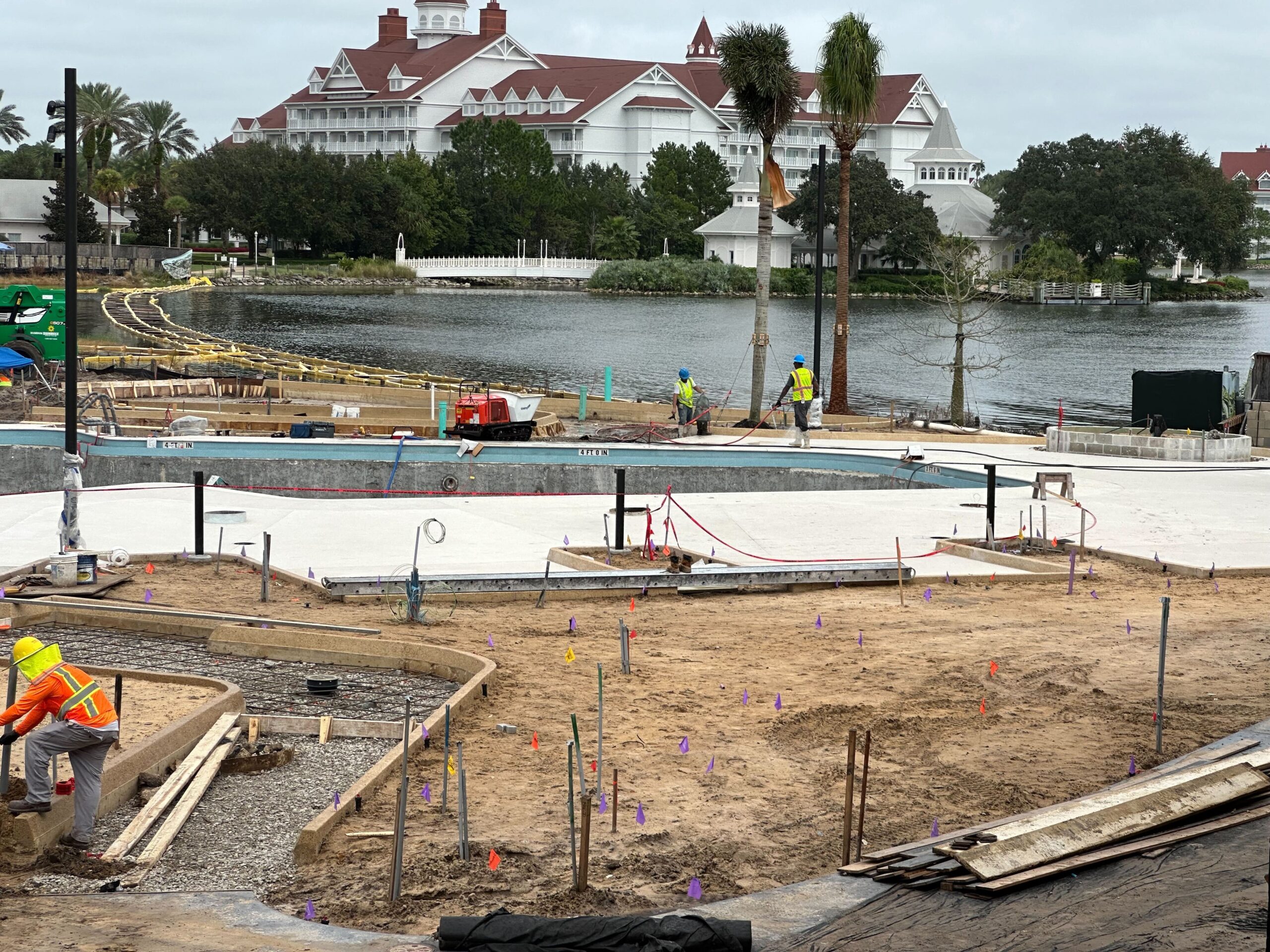 Construction workers are building a concrete pathway near a waterfront, with a large building featuring red roofs and white walls in the background.