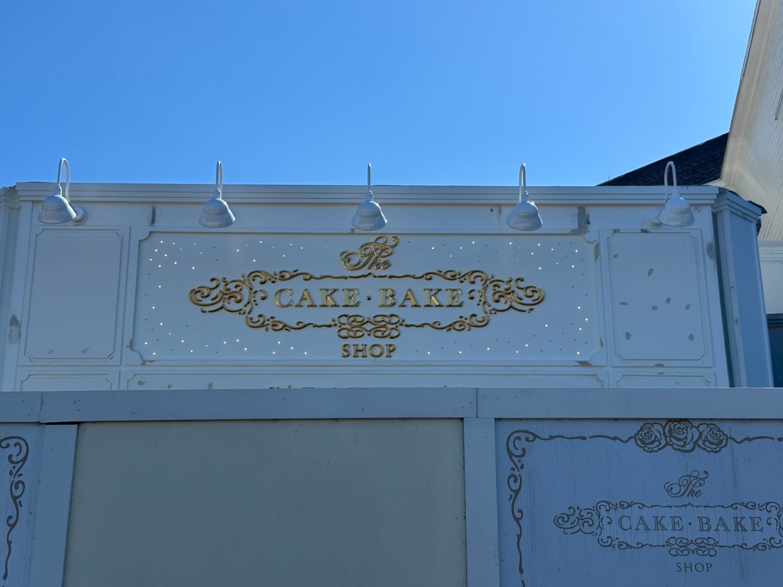 A light blue and white Cake Bake Shop storefront sign with gold lettering is illuminated by four lights under a clear blue sky.