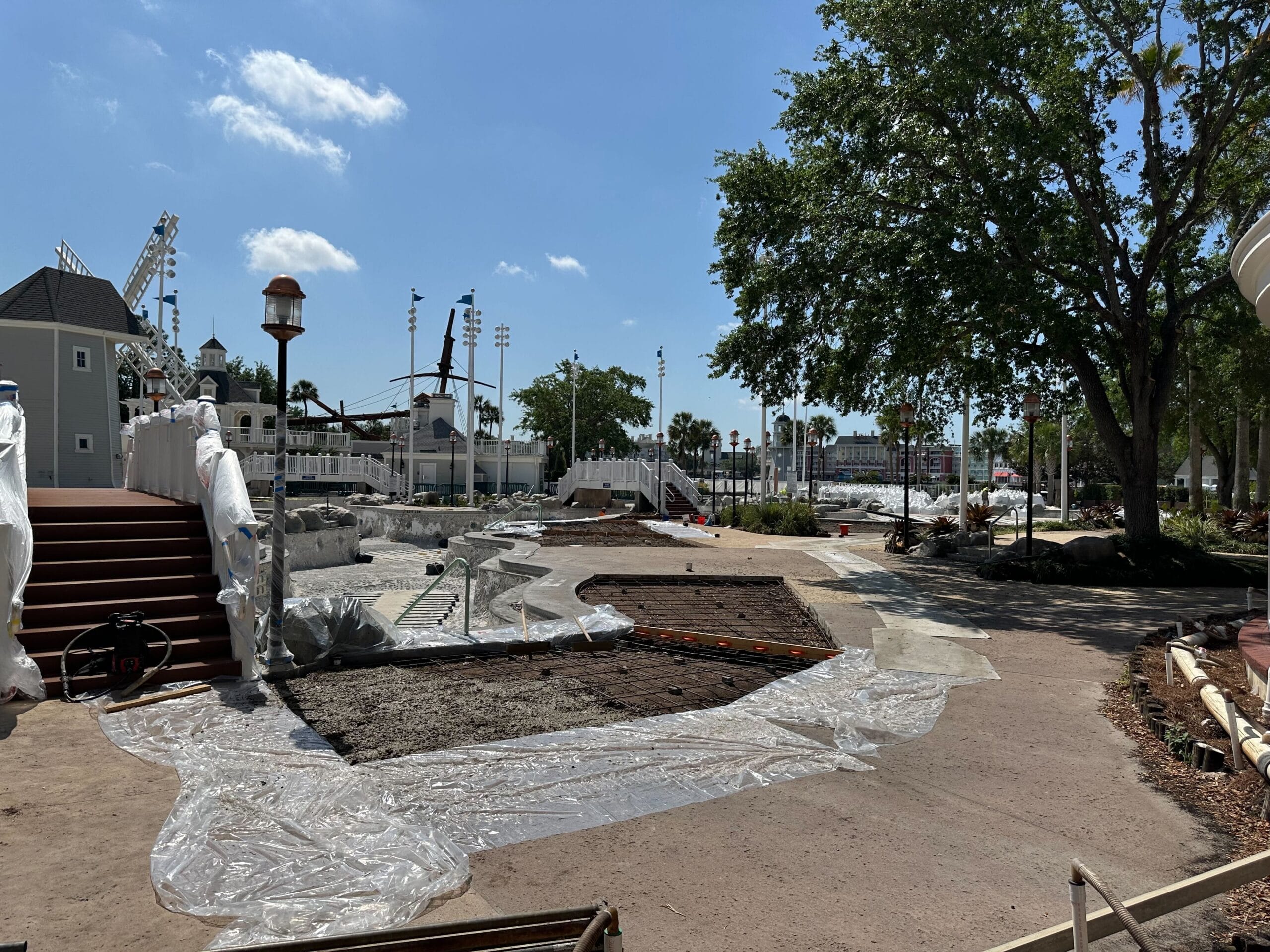 The construction site features a tower of new scaffolding amid partially built structures, plastic coverings, and scattered materials. Trees frame the scene under a clear blue sky, offering a view reminiscent of Disney’s Yacht Club's serene surroundings.