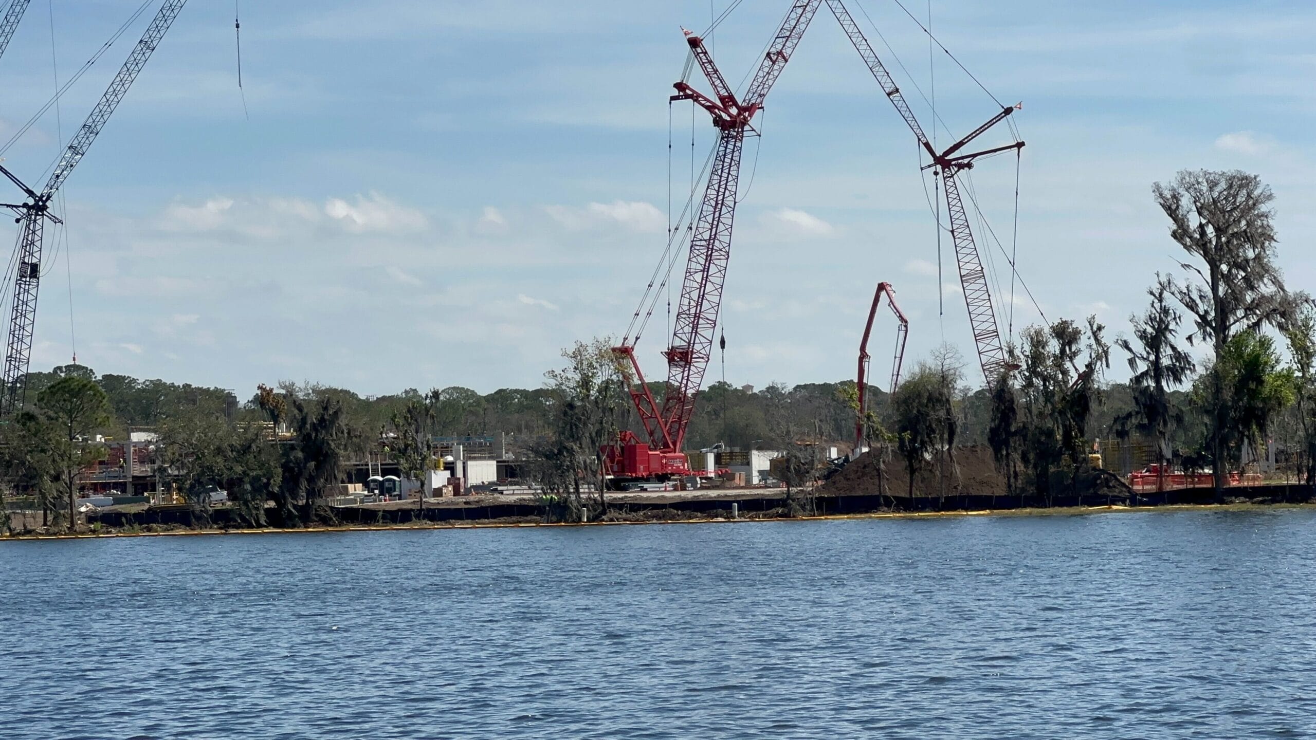 The construction site for the Lakeshore Lodge is bustling with several cranes by the lake, trees standing tall in the background, and a clear blue sky above.