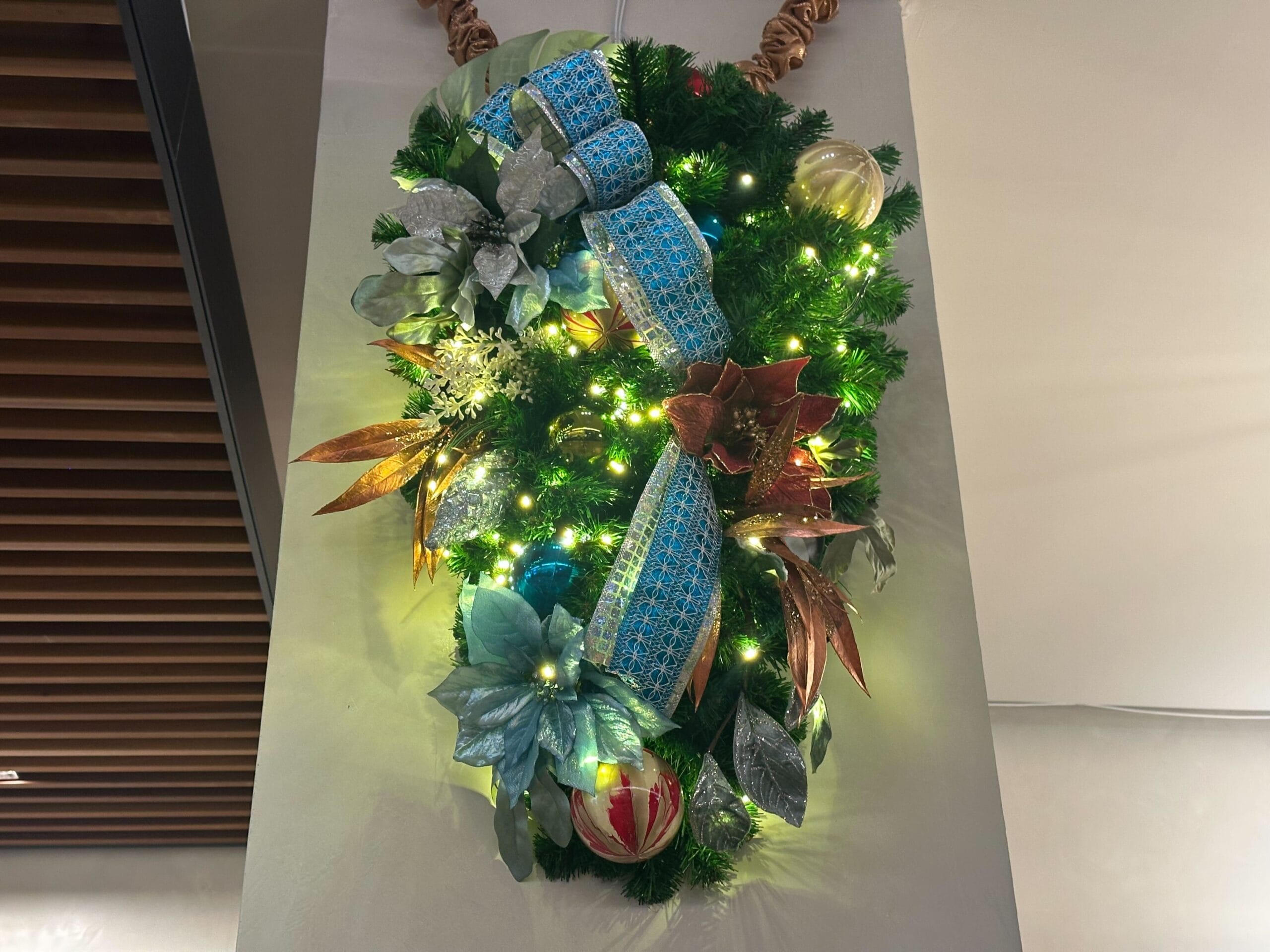 Christmas wreath with glowing lights, blue ribbon, and decorative flowers and ornaments hangs on a wall.