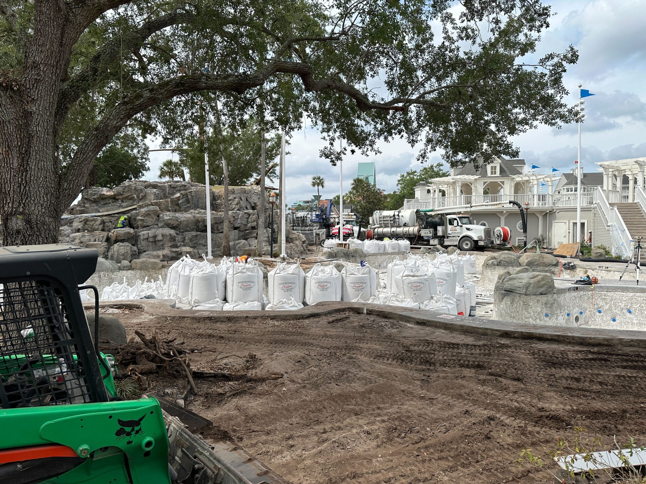 A construction site with dirt, large white sandbags, trucks, and workers near a white building and rocky area under a tree.