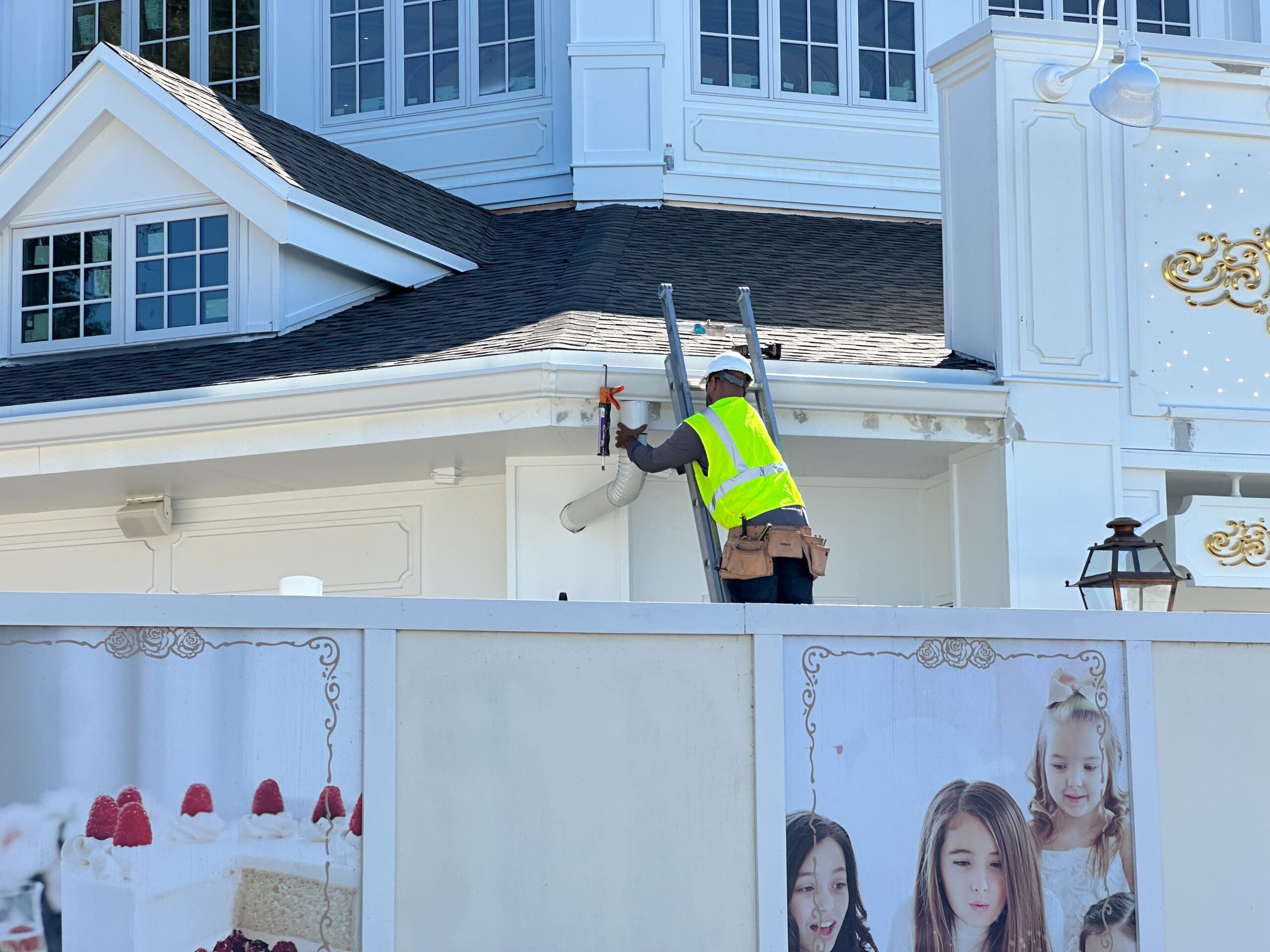 Worker in a fluorescent vest on a ladder fixing the exterior of a building, with a decorative fence featuring photos of people and desserts in the foreground.