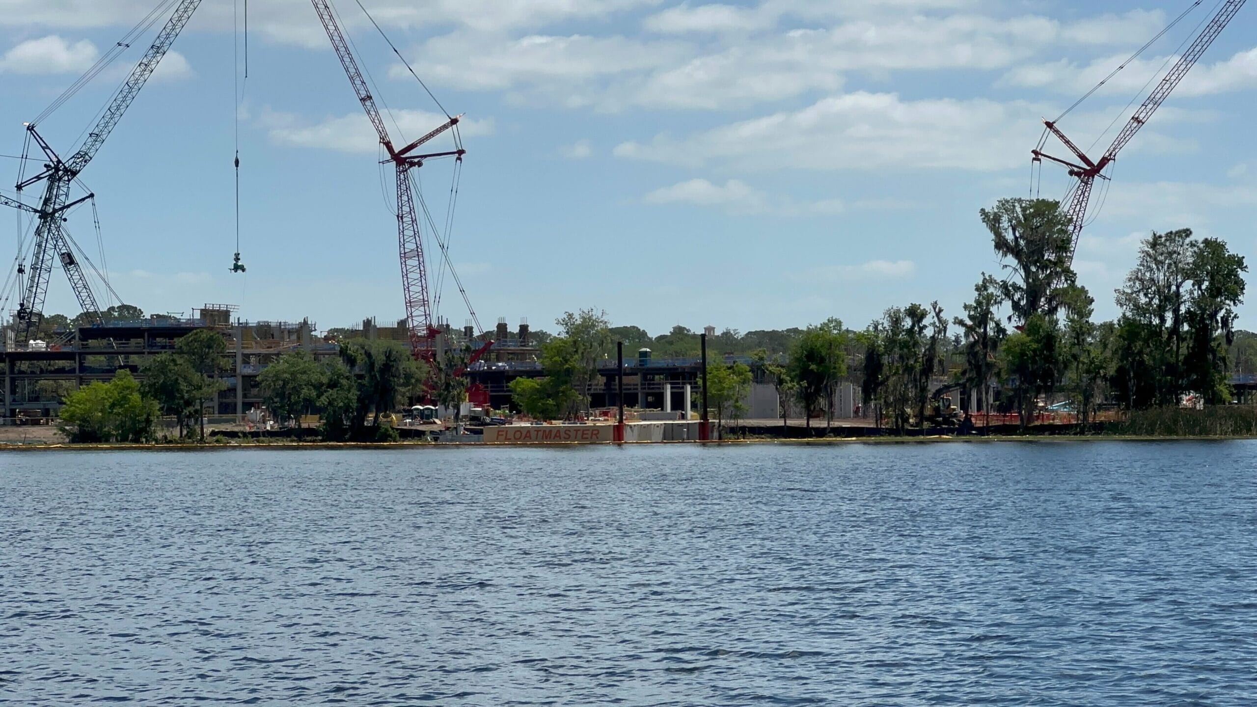 Construction site with cranes beside a waterfront, trees lining the water's edge, and buildings in the background under a partly cloudy sky.