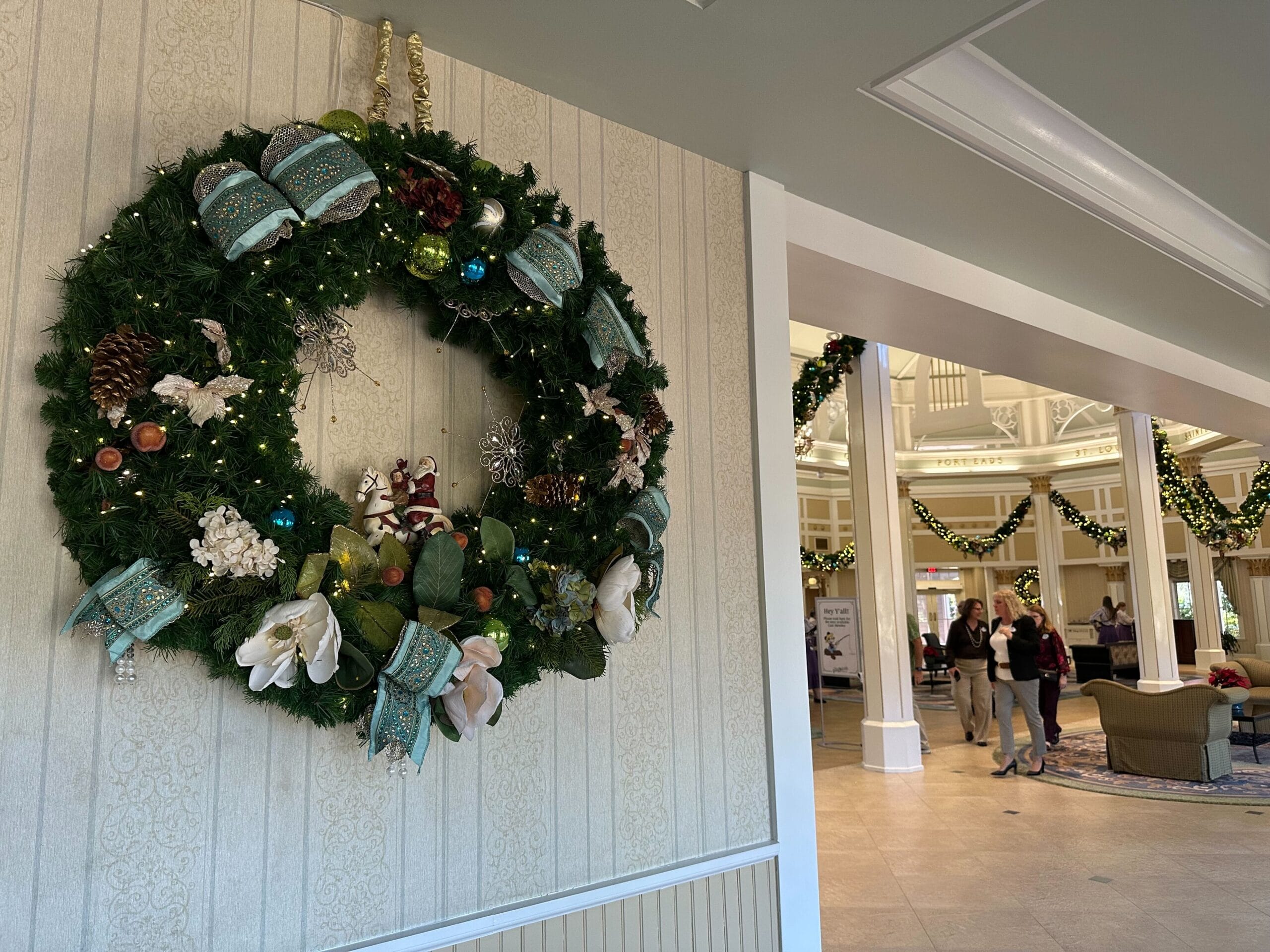 Large holiday wreath with ornaments and bows on a wall. Lobby area in the background decorated with garlands, people walking, and seating arrangements visible.