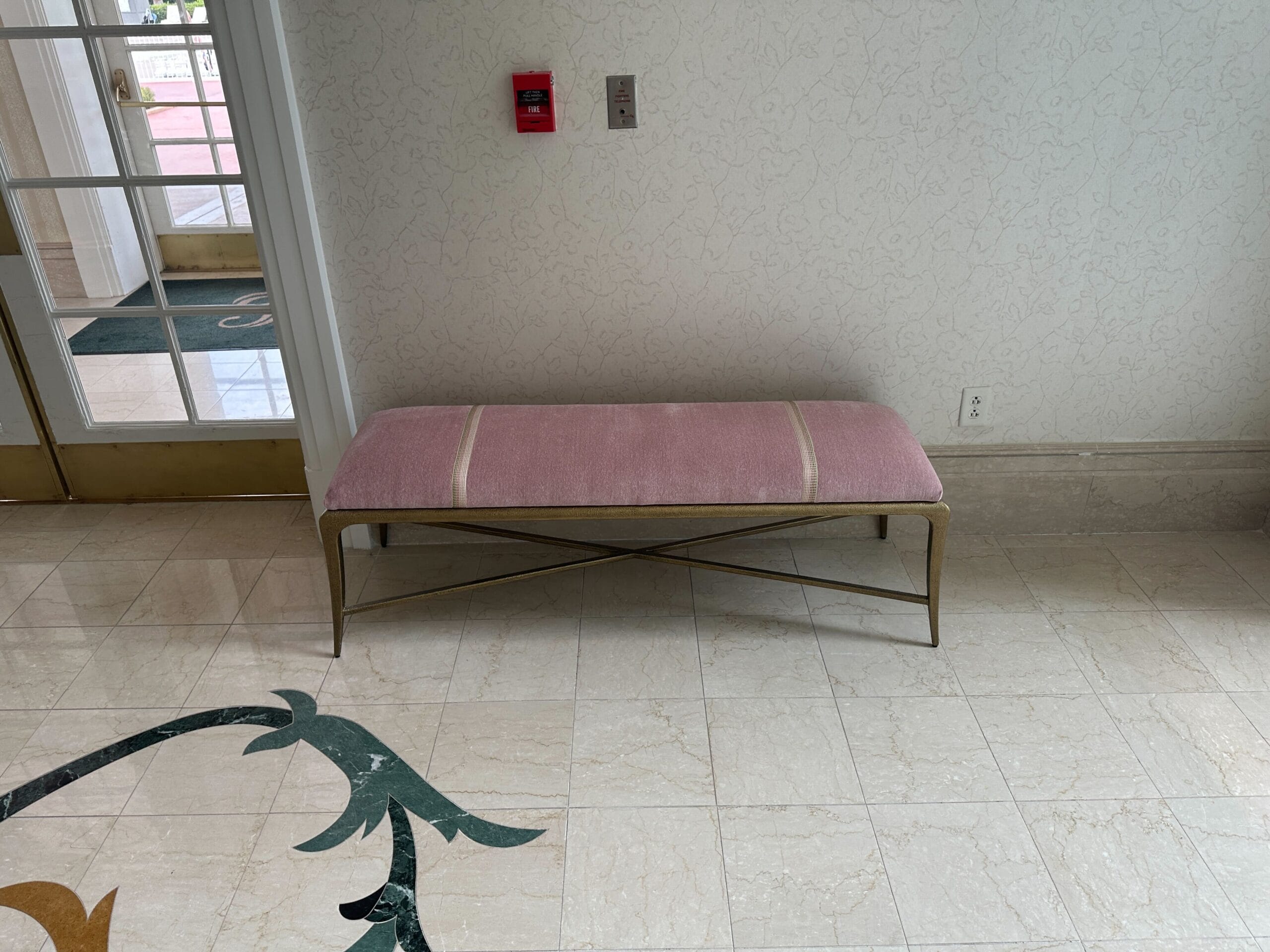A pink cushioned bench with gold legs sits against a wall in a tiled hallway near a door.
