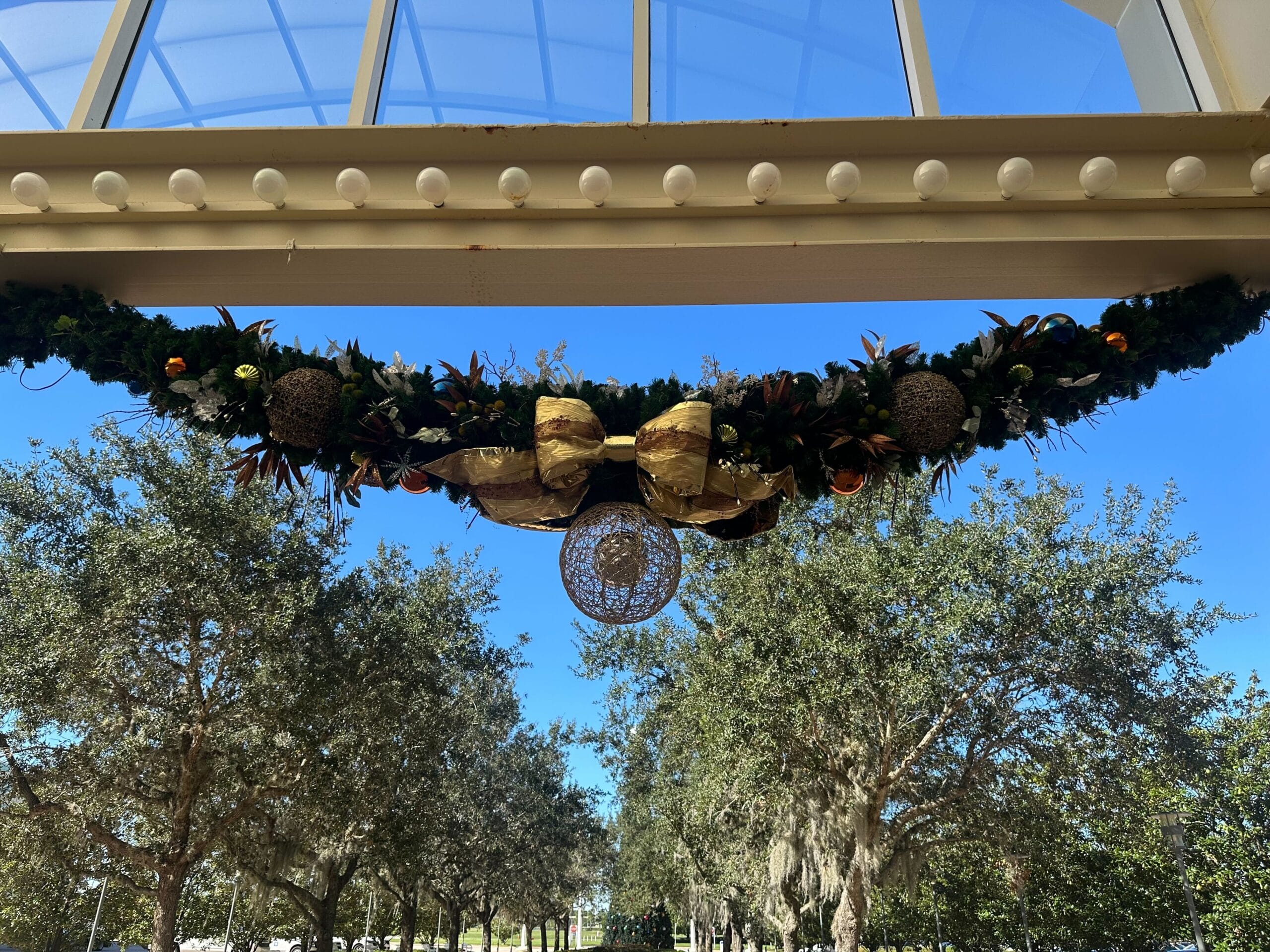 Garland with gold bows and ornaments hanging under a glass ceiling with trees and a clear blue sky in the background.