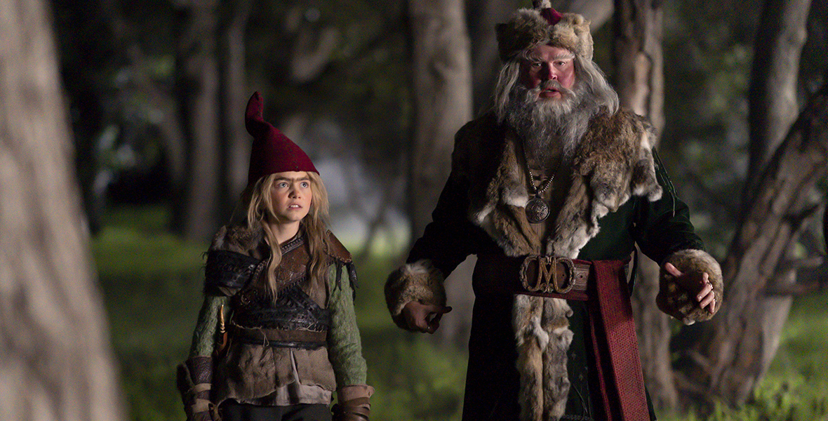 In an image from The Santa Clauses Season 2,  Magnus Antas (Eric Stonestreet) is dressed in a brown coat with a fur rim, gray pants, a fur hat, and brown boots; he has a grey beard and long hair. He’s standing next to a boy wearing an elf hat, green top, and black pants. The pair is standing in a forest.