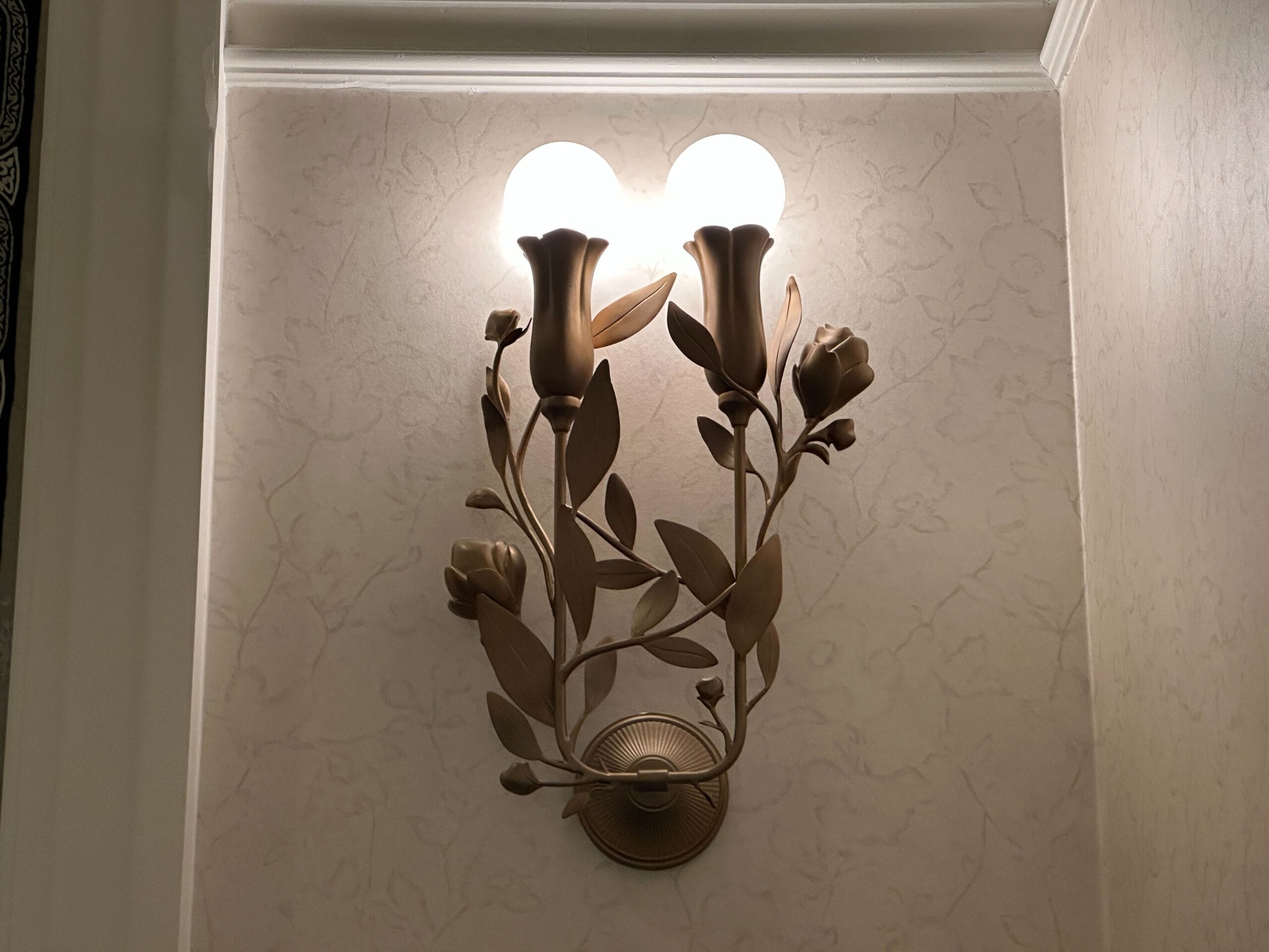 Wall sconce with two lit bulbs and Grand Floridian-inspired golden floral accents on a textured beige wall.